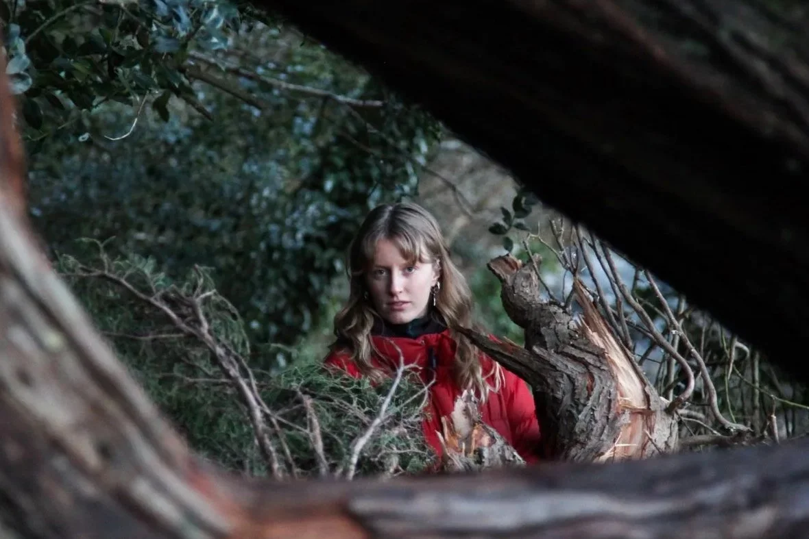 A young woman with wavy blonde hair wearing a red jacket and earrings, looking through a natural circular opening in a fallen tree surrounded by green foliage.