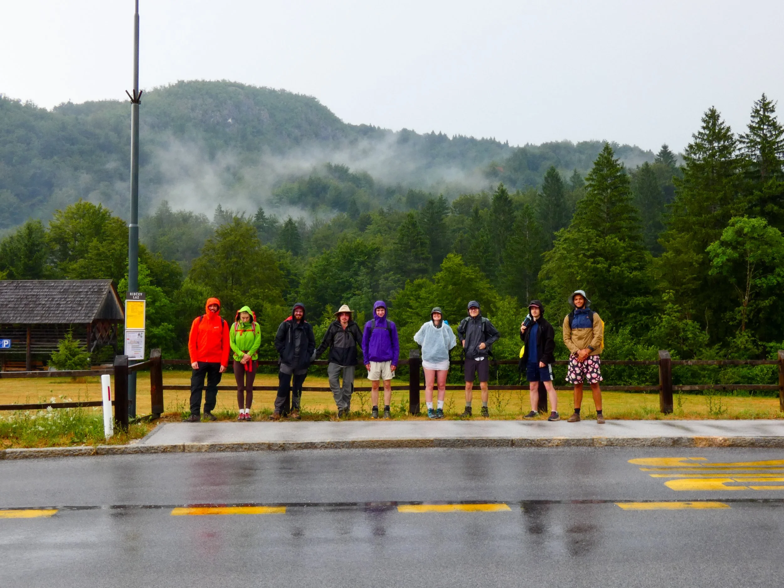 A group of nine people standing in a line on a sidewalk in front of a wooden fence, dressed in rain gear, with trees and foggy mountains in the background on a rainy day.