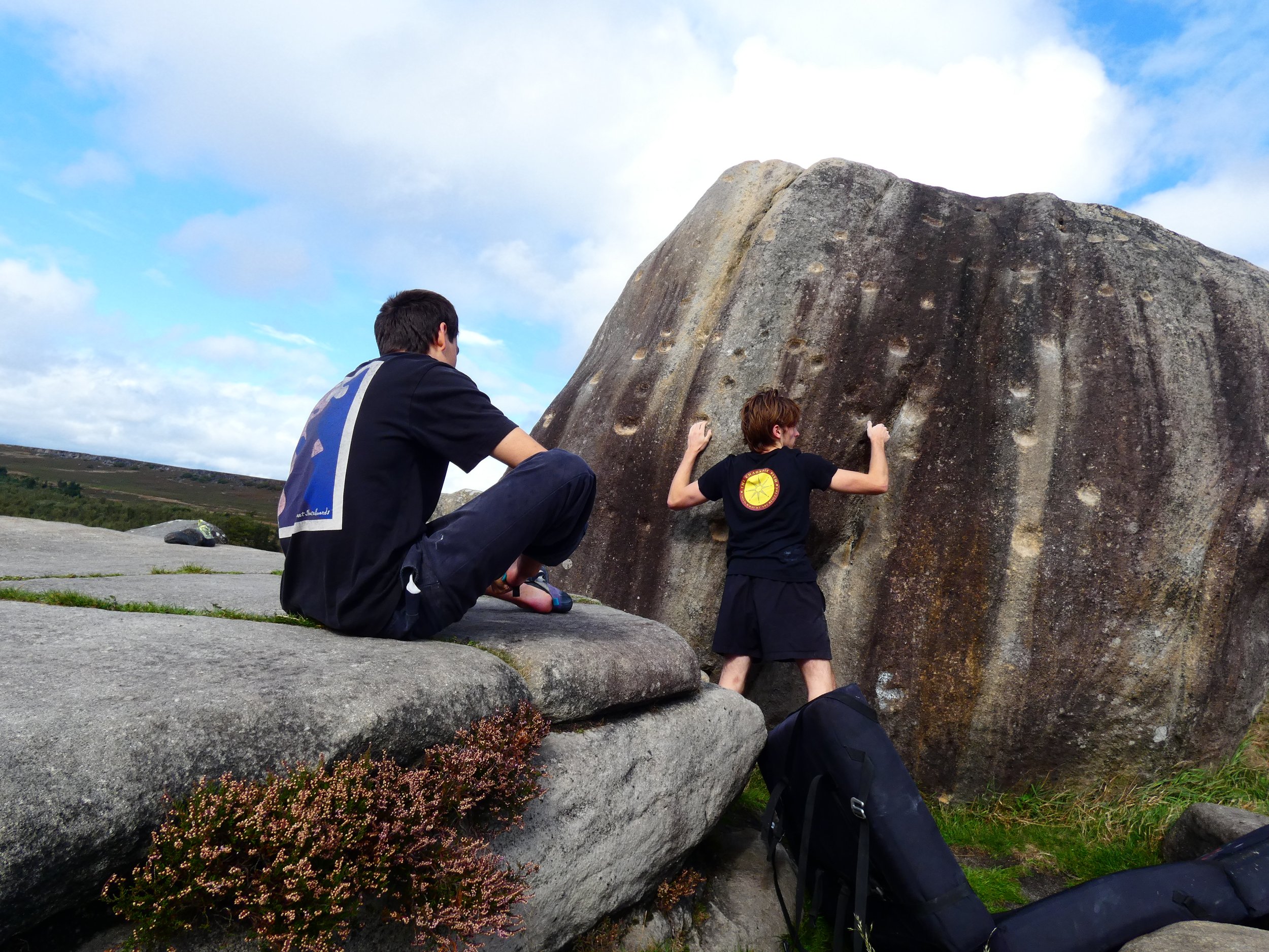 Two young climbers on a rocky outdoor terrain, with one sitting and another climbing a large boulder under a partly cloudy sky.