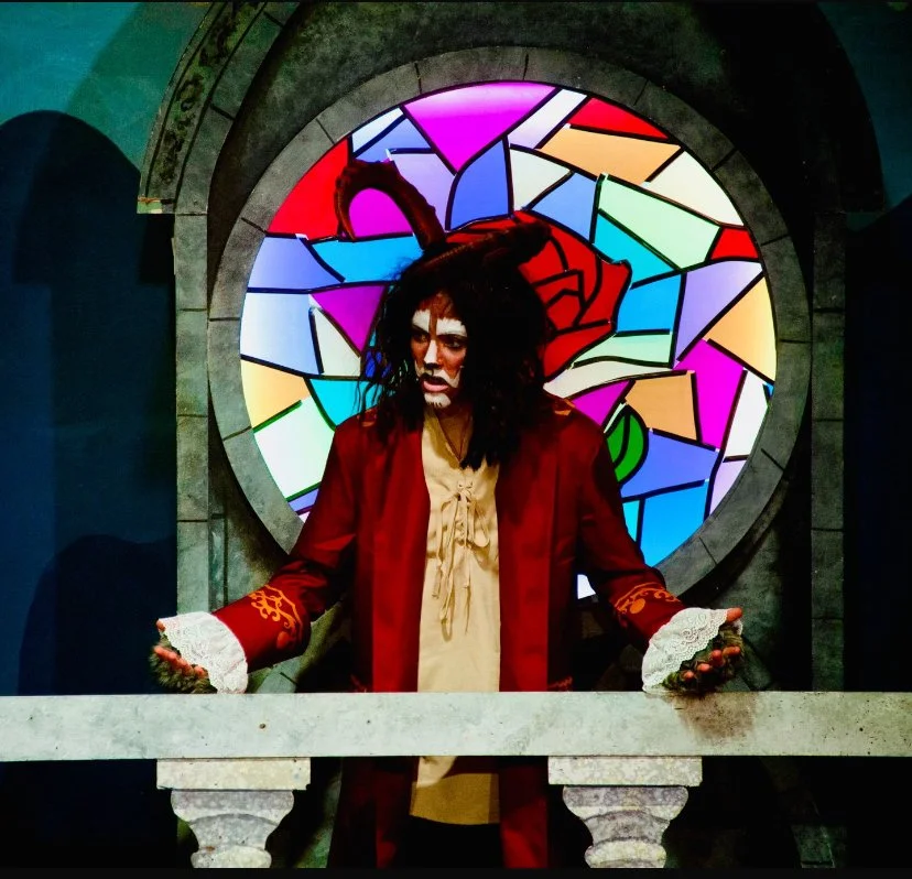Costumed character with dark, messy hair and animal-like ears, dressed in a red coat with lace cuffs, standing in front of a stained glass window with an abstract design and a rose image, inside a stone architectural setting.