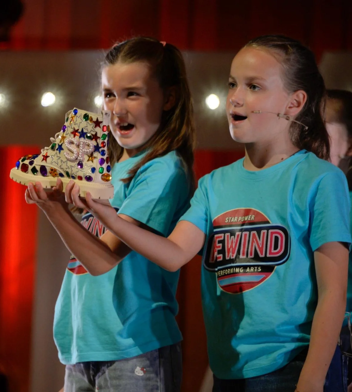 Two young girls with long hair and light blue T-shirts on stage, one holding a decorated rubber boot with colorful gems and the word 'SP' on it, appearing surprised or excited.