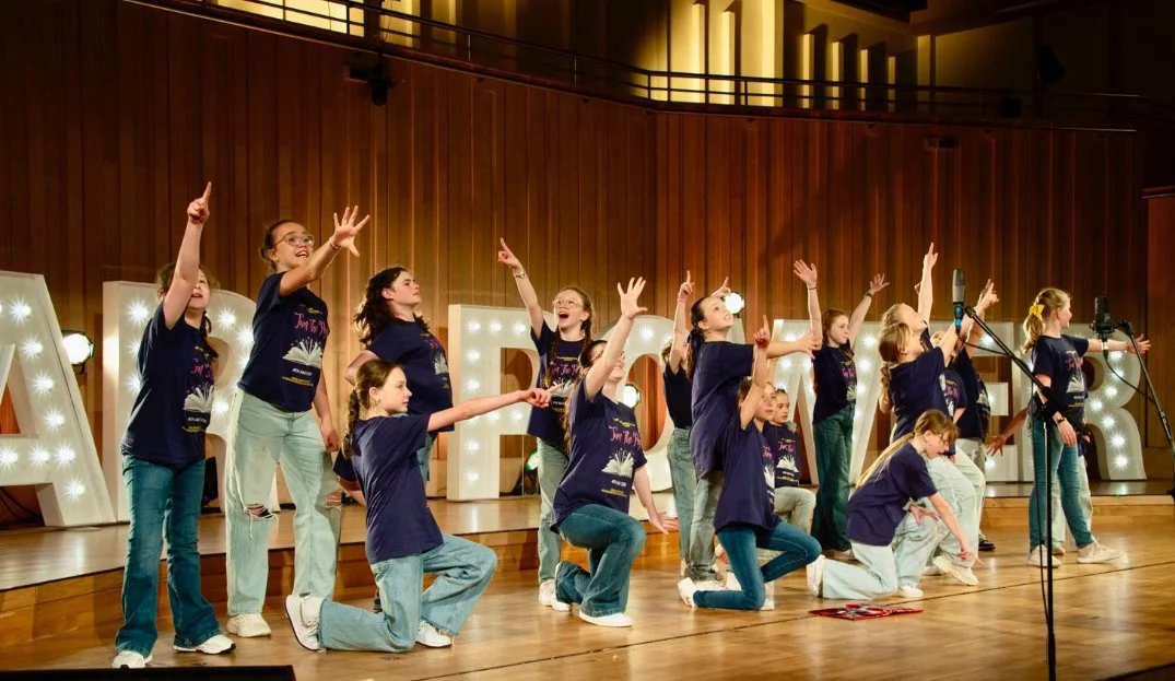 Group of young girls performing on stage with large illuminated letters behind them, wearing matching navy blue t-shirts.
