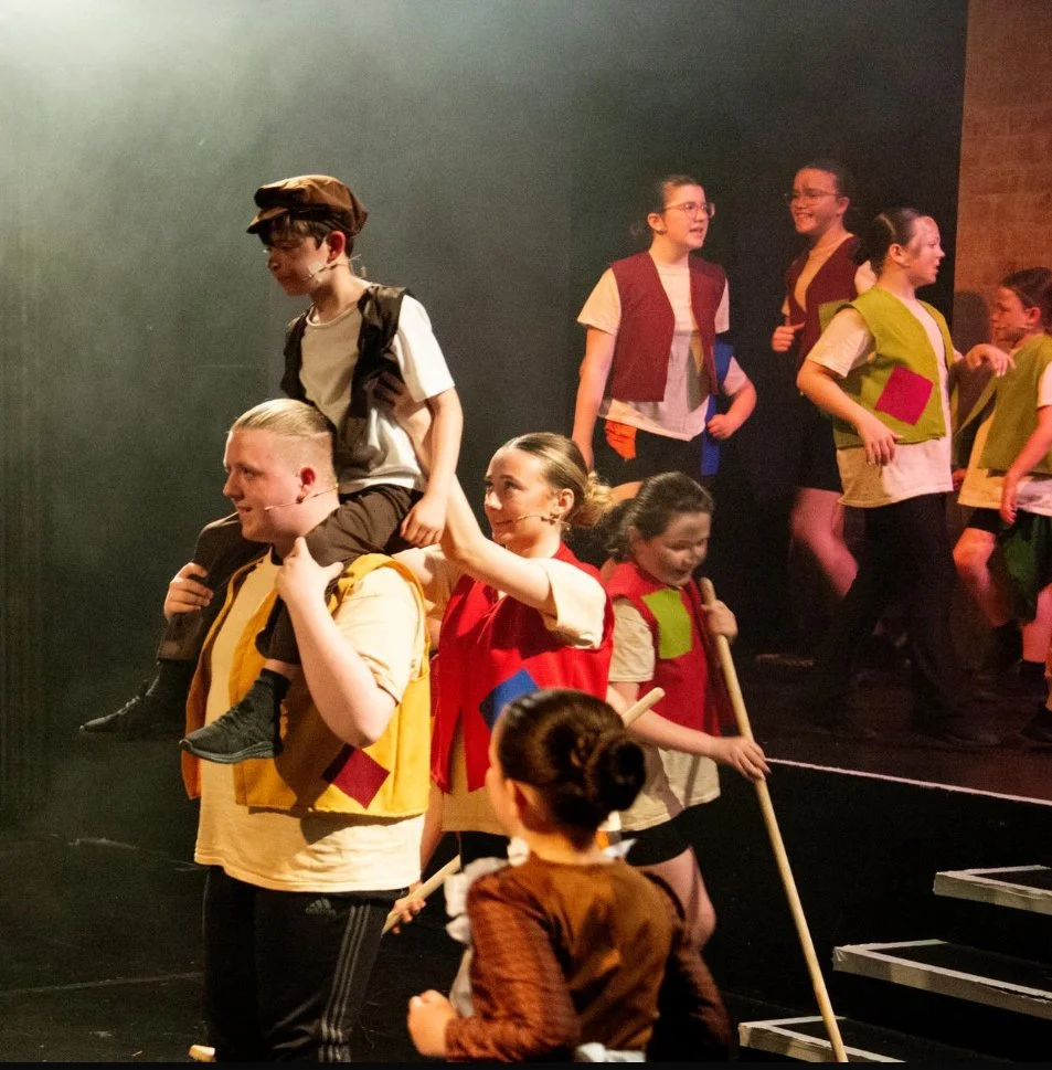 Children are performing on stage during a play or rehearsal, with some wearing costumes and holding props.