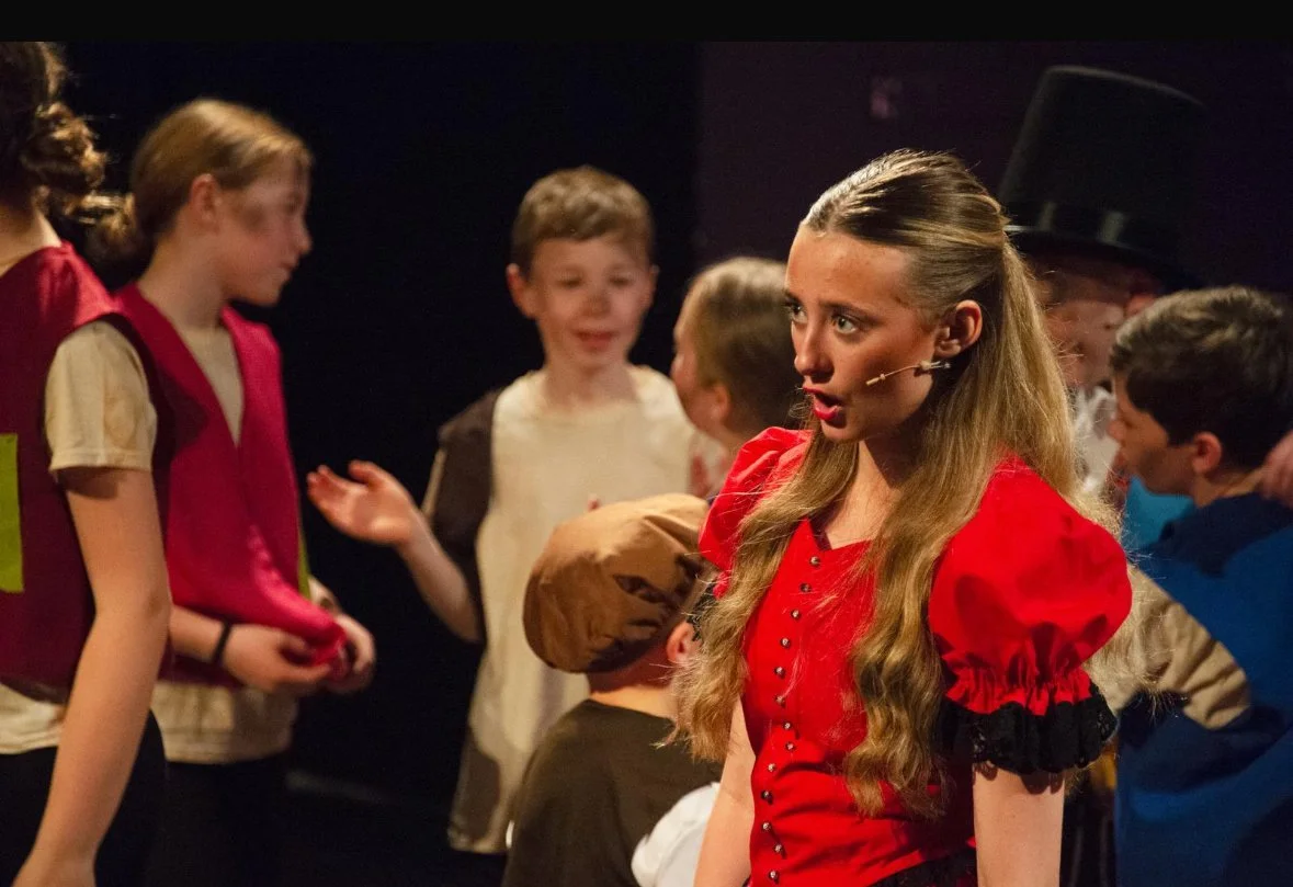 Children on stage during a theatrical performance, with a young girl in red costume in the foreground, speaking into a headset microphone.