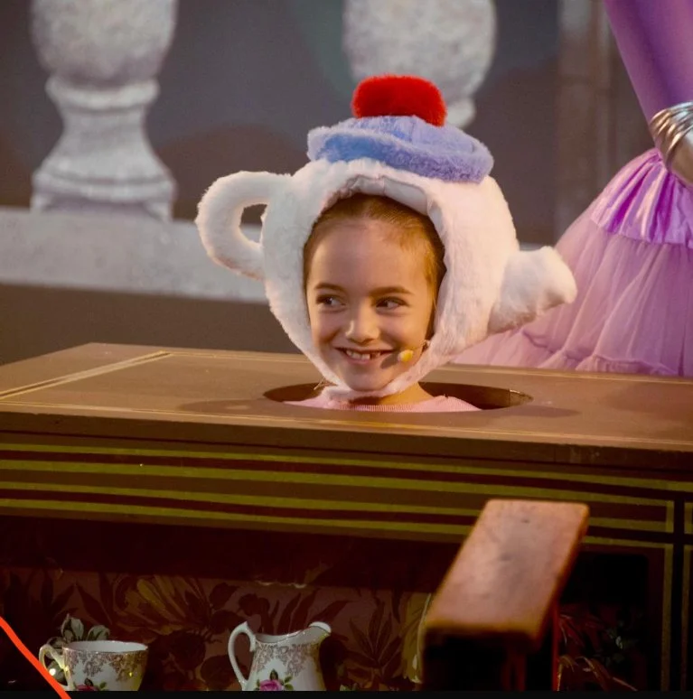 A young girl in an elaborate Teapot costume with a fluffy white hat resembling a teapot lid, a red pom-pom, and a purple lid, participating in a game where her head is popping out of a hole in a cardboard prop.