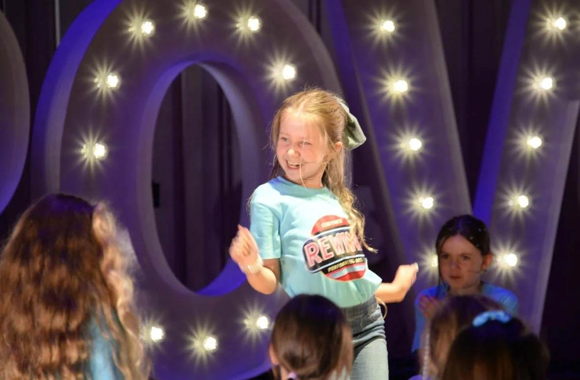 A group of children, including a girl with a large bow in her hair, on stage with illuminated decorations in the background. The girl is smiling and appears to be performing or speaking.