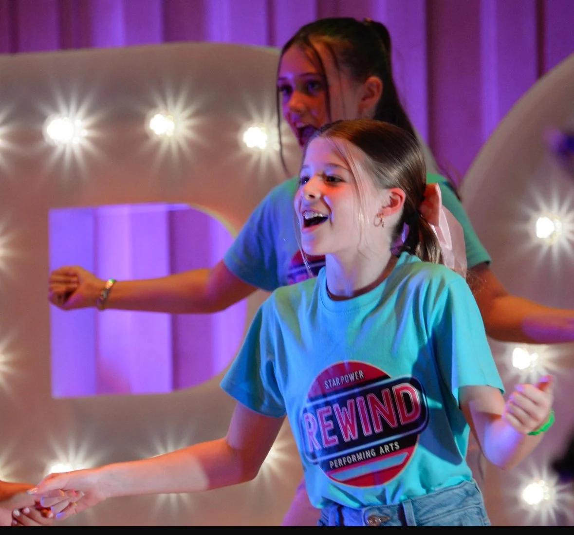 Two young girls smiling and dancing in front of a decorative backdrop with lights. The girl in the foreground wears a light blue T-shirt with a 'Rewind' logo.