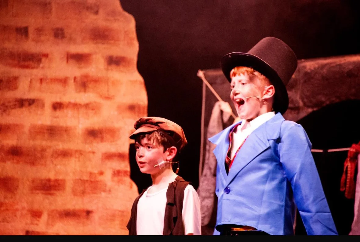 Two boys dressed as characters from a theatrical play, with one wearing a tall black hat, a blue jacket, and a white shirt, and the other wearing a cap and a vest, standing on stage in front of a brick background.