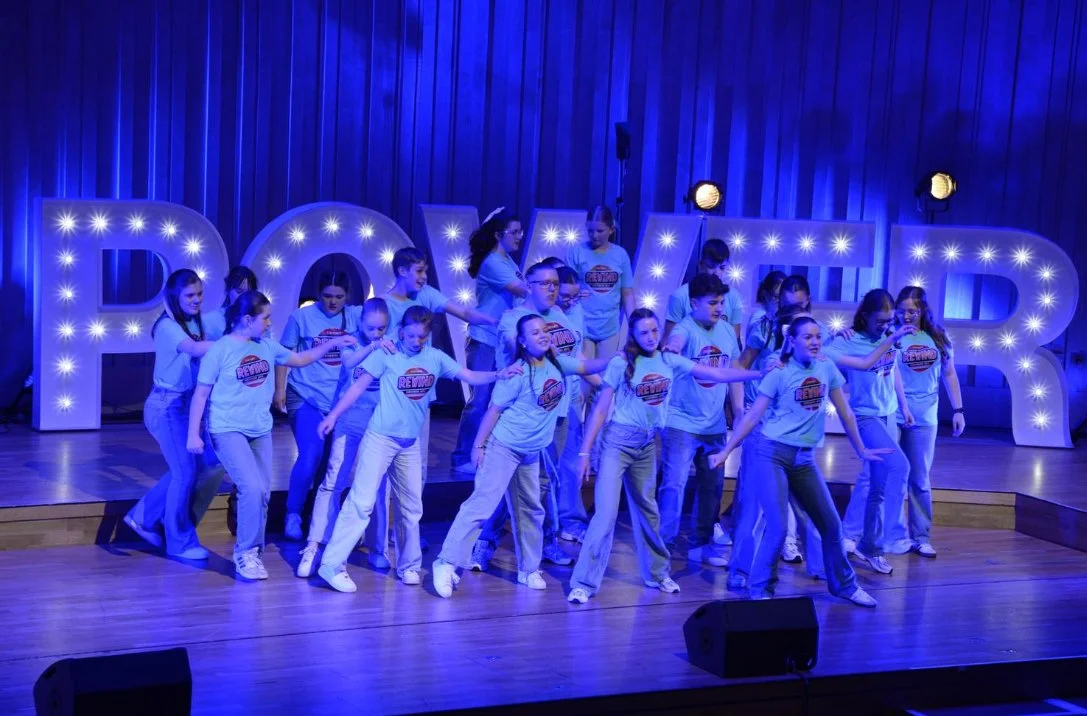 Group of children performing dance on stage with large illuminated letters spelling 'REWARD' in the background.