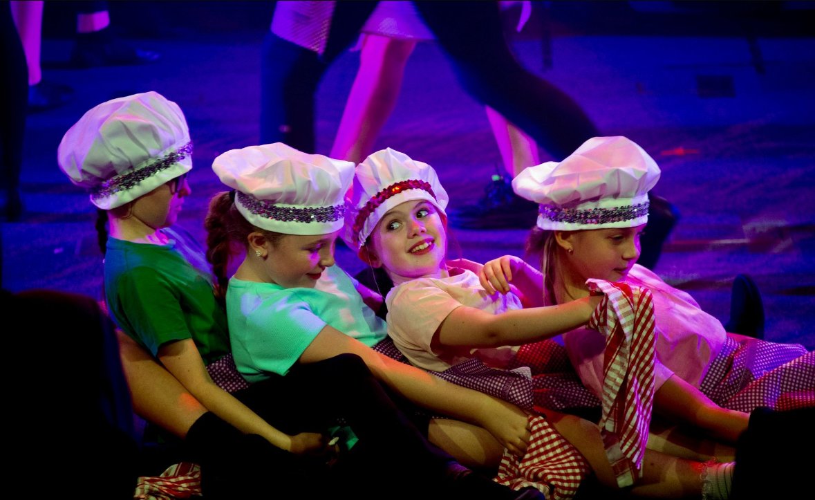 Four young girls dressed as chefs with white chef hats and colorful costumes sitting on stage, with purple and blue lighting, during a performance.