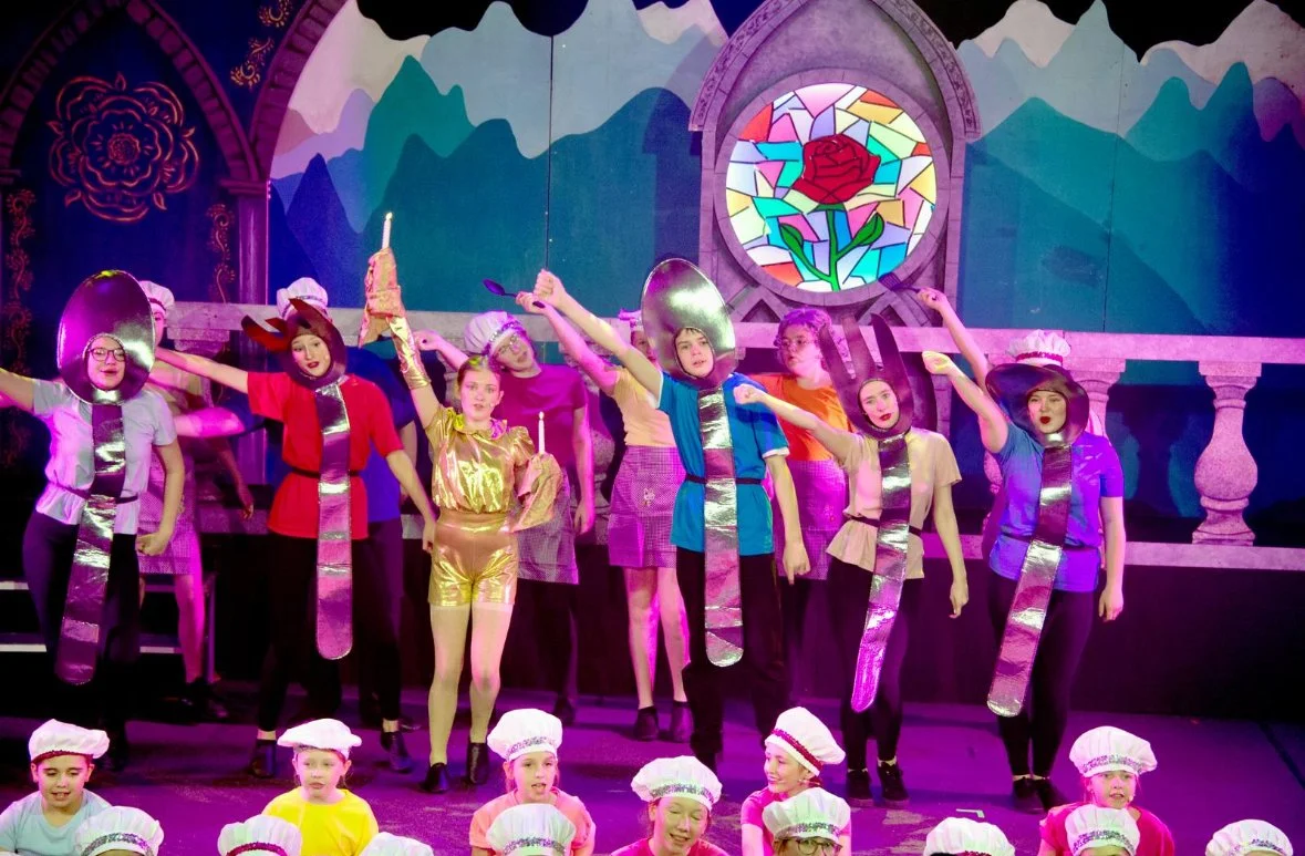 Children performing a theatrical play on stage, dressed in colorful costumes with shiny accessories, in front of a backdrop with mountain scenery and a stained glass window featuring a red rose.