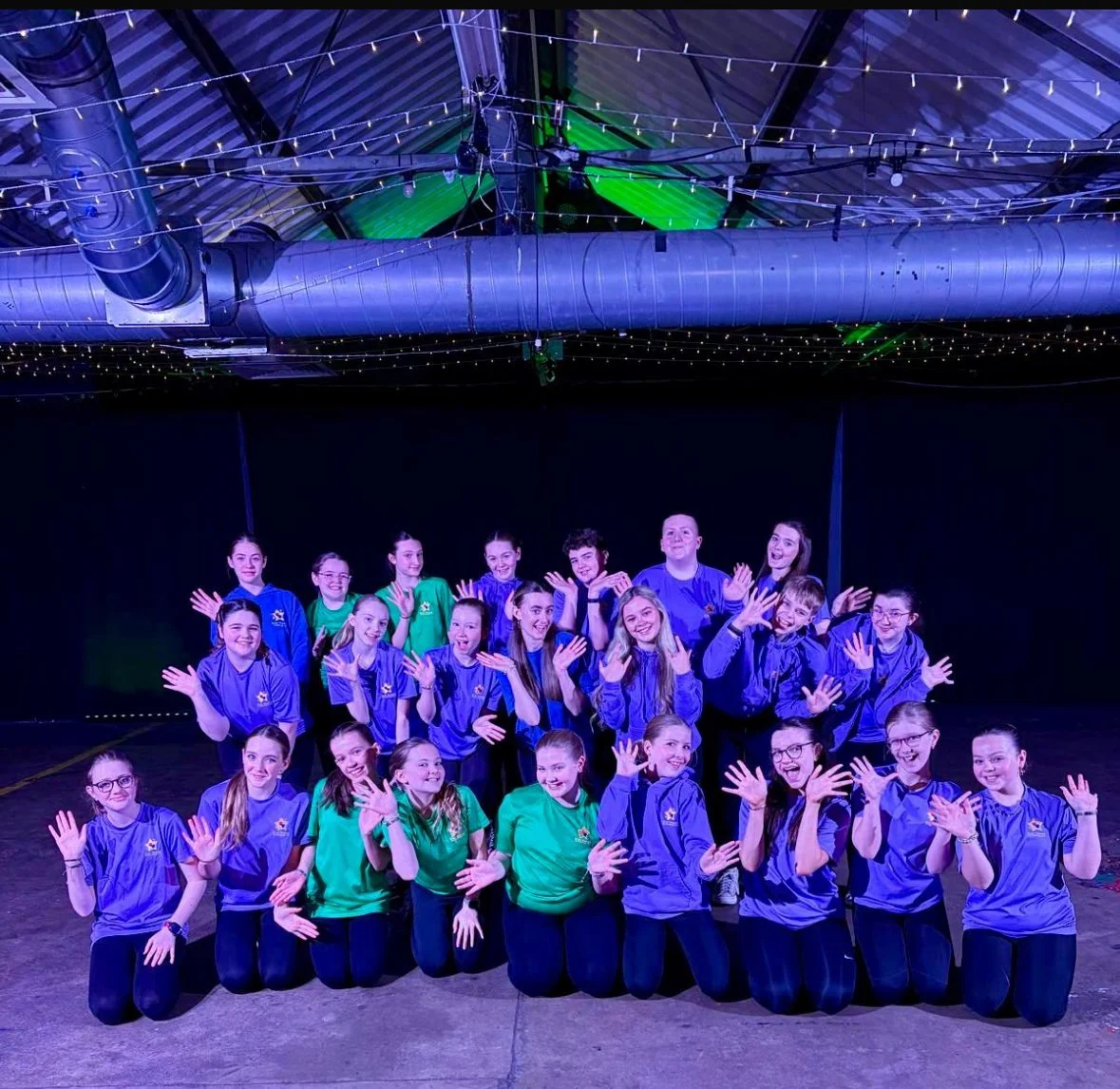 Group of young girls and boys in colorful sports uniforms posing together on stage with their hands raised in a friendly gesture, under string lights and industrial ceiling pipes.