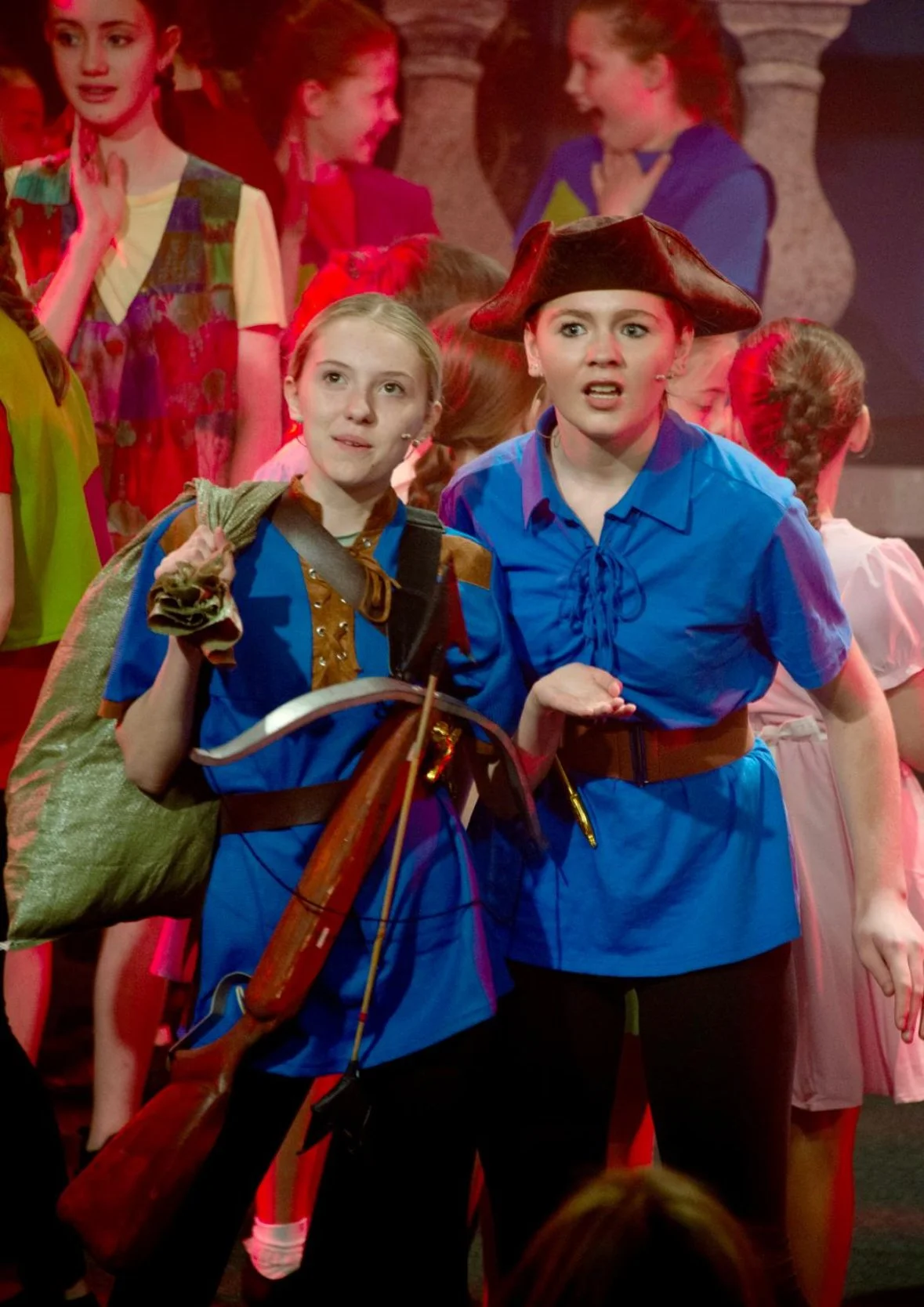 Two young women dressed in costumes on stage, surrounded by other girls. One is wearing a brown and blue costume with a bow and arrow, and the other in a blue dress with a pirate hat, both appearing surprised or confused.