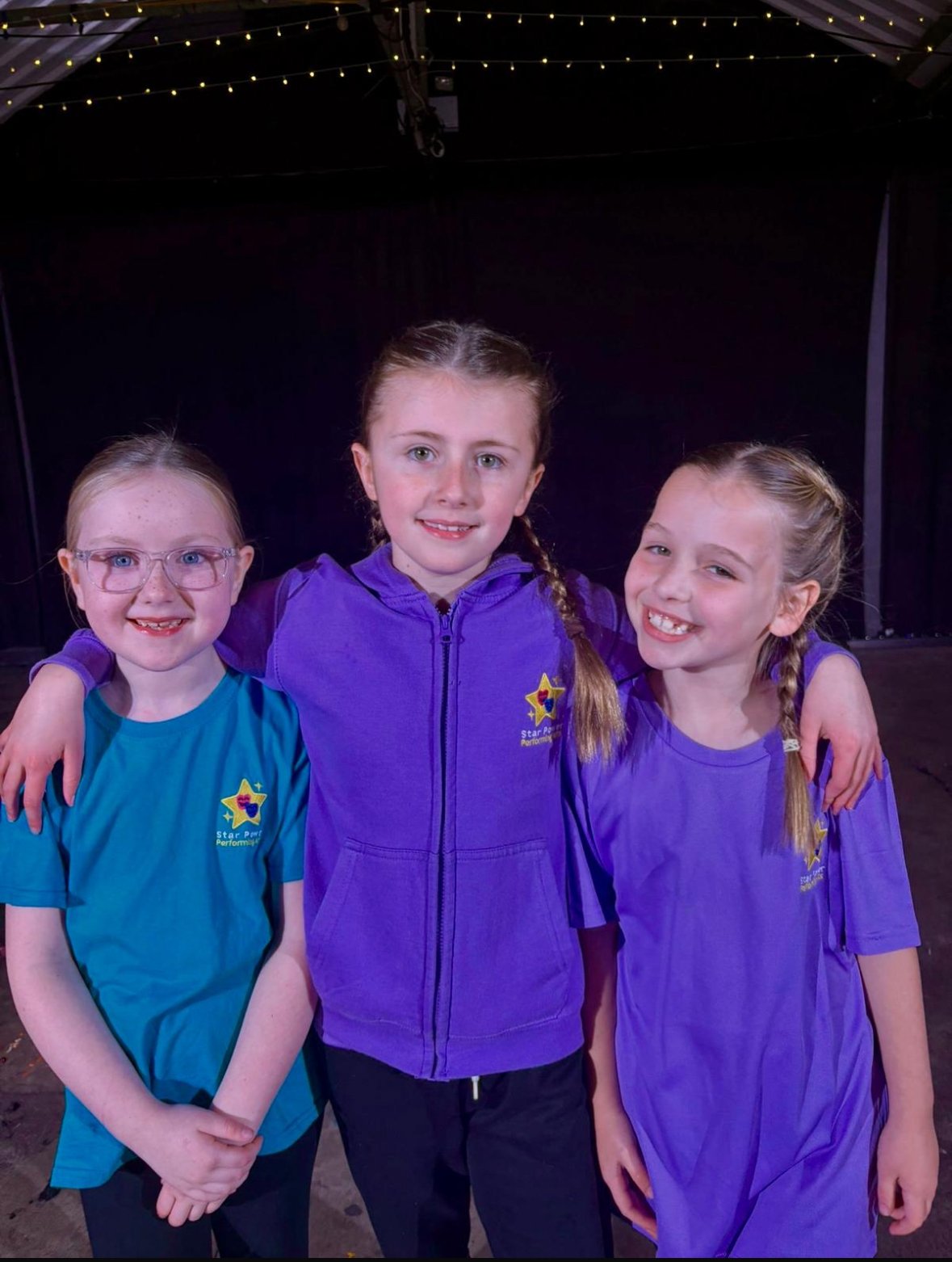 Three young girls standing together on a stage, smiling, wearing colorful performance shirts with a star logo, with string lights hanging above them.