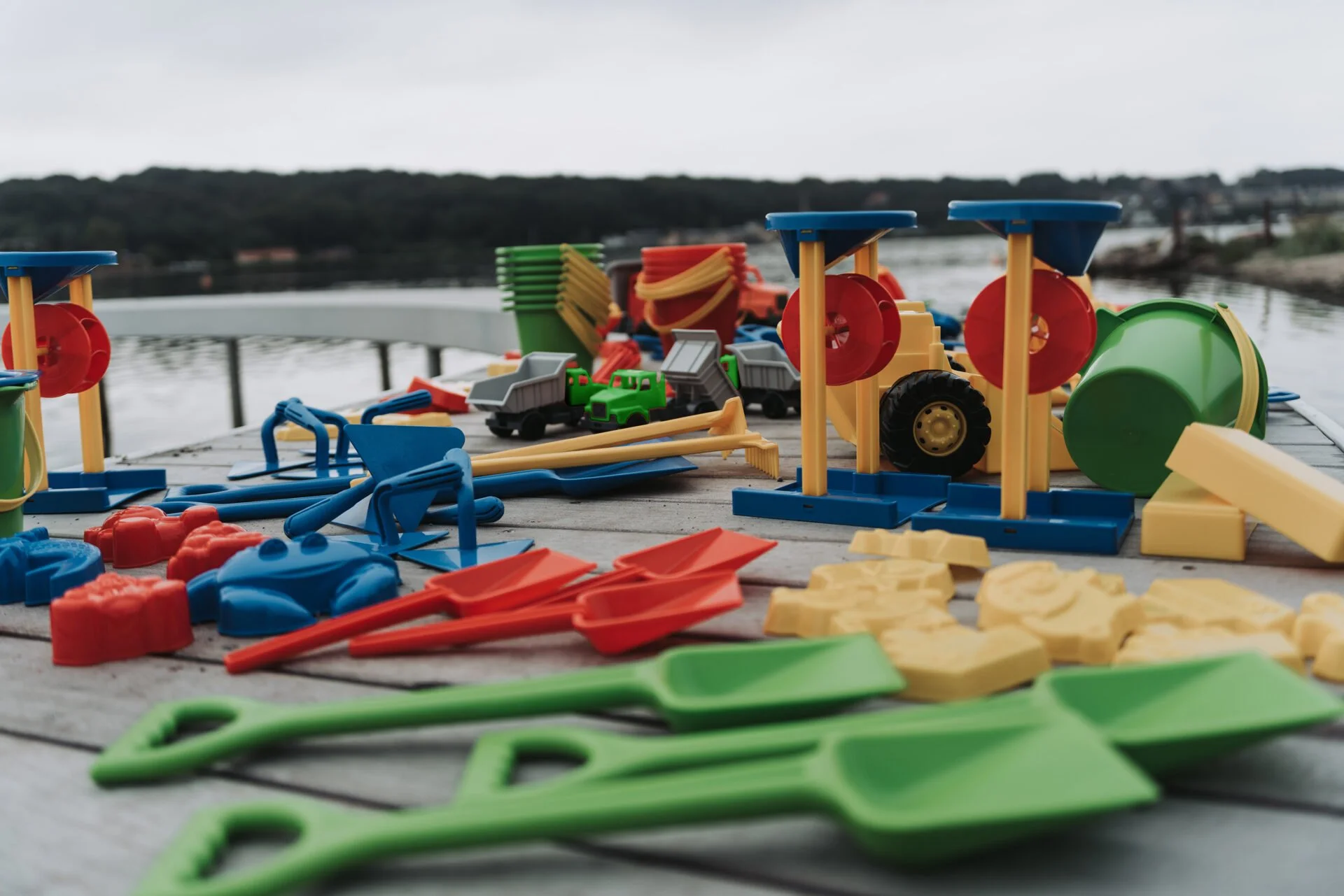 Colorful toy construction and transportation playset including shovels, rakes, dump trucks, and buckets on a wooden dock overlooking a body of water.