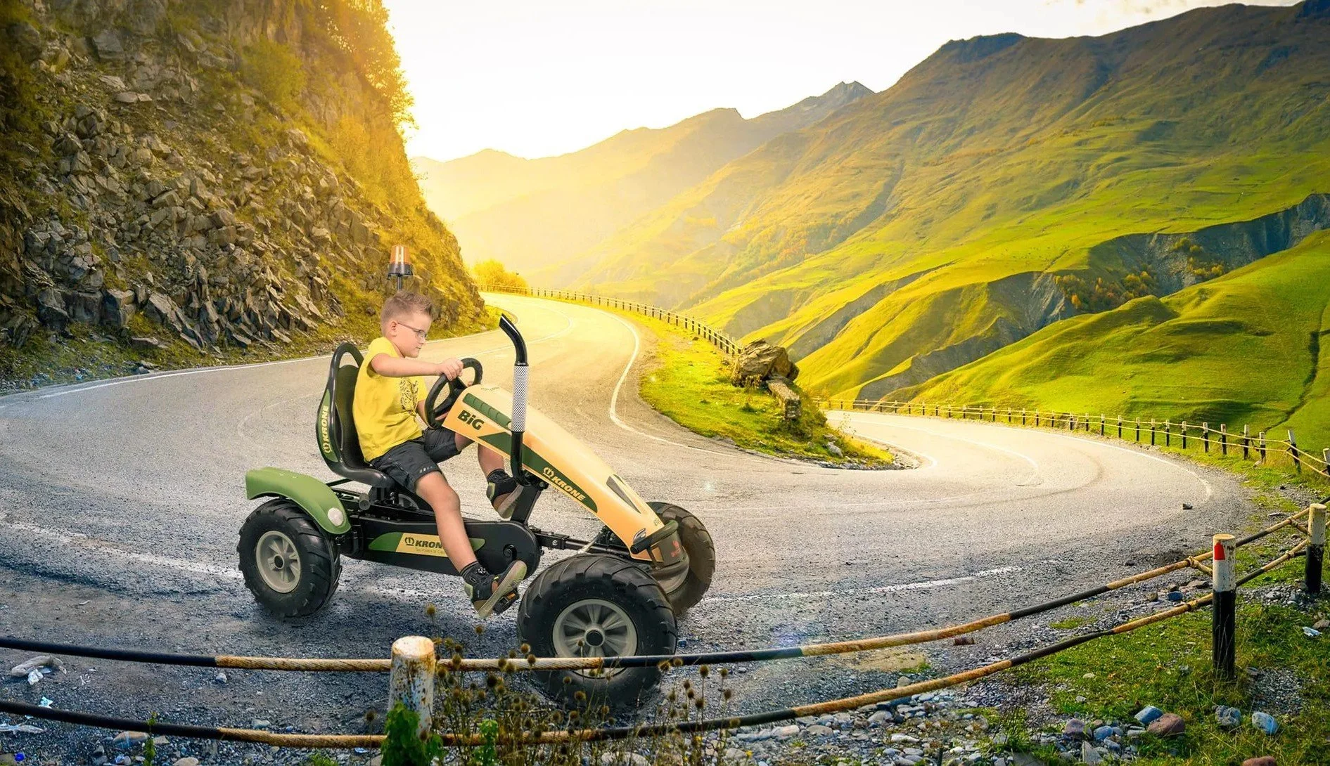 A child riding a toy ride-on quad bike on a winding mountain road with green hills and mountains in the background during sunset.