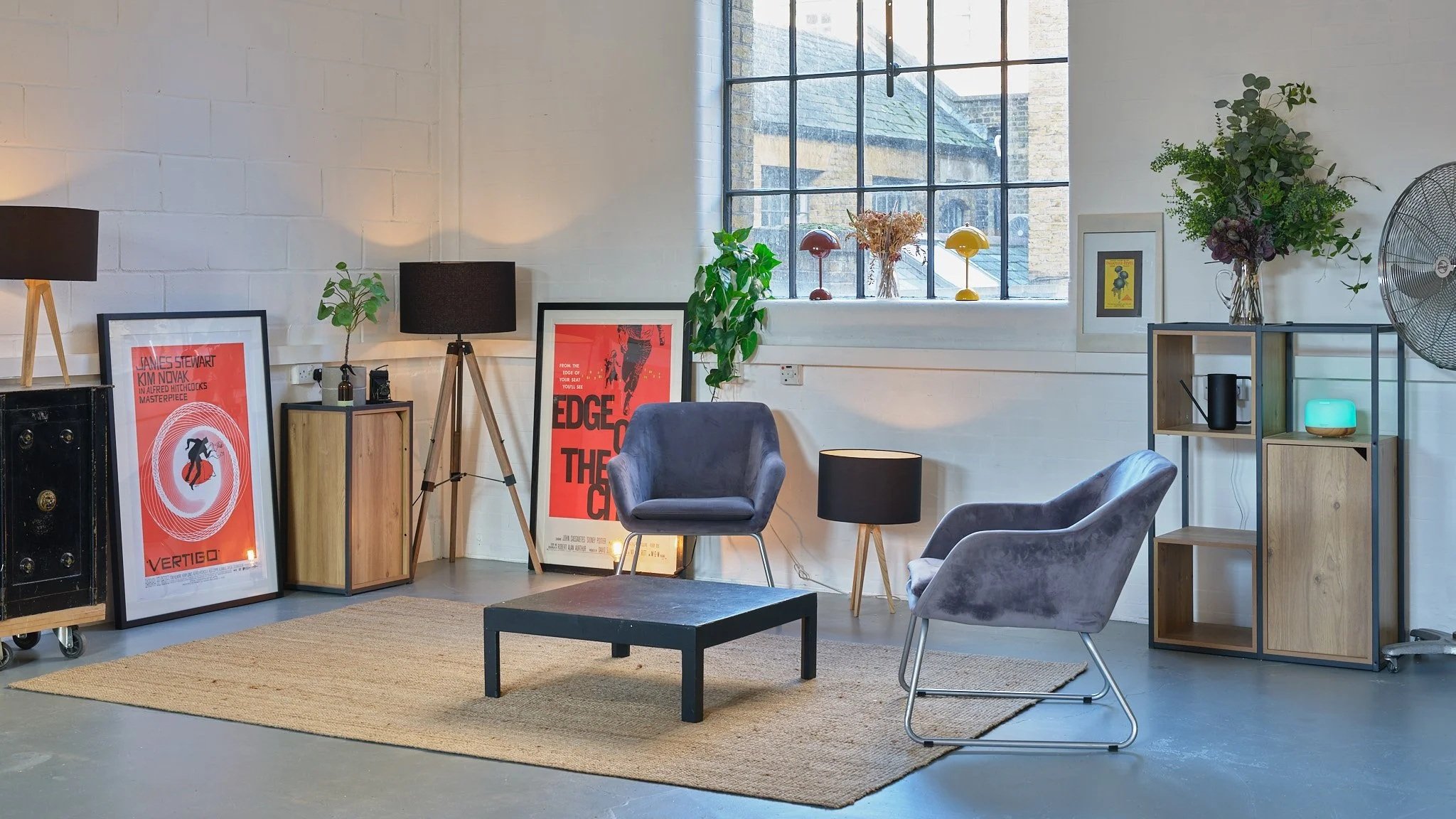 Modern living room with two gray chairs, a black coffee table, a beige rug, and assorted framed posters and decorative items on shelves and walls, illuminated by natural light from a large window.