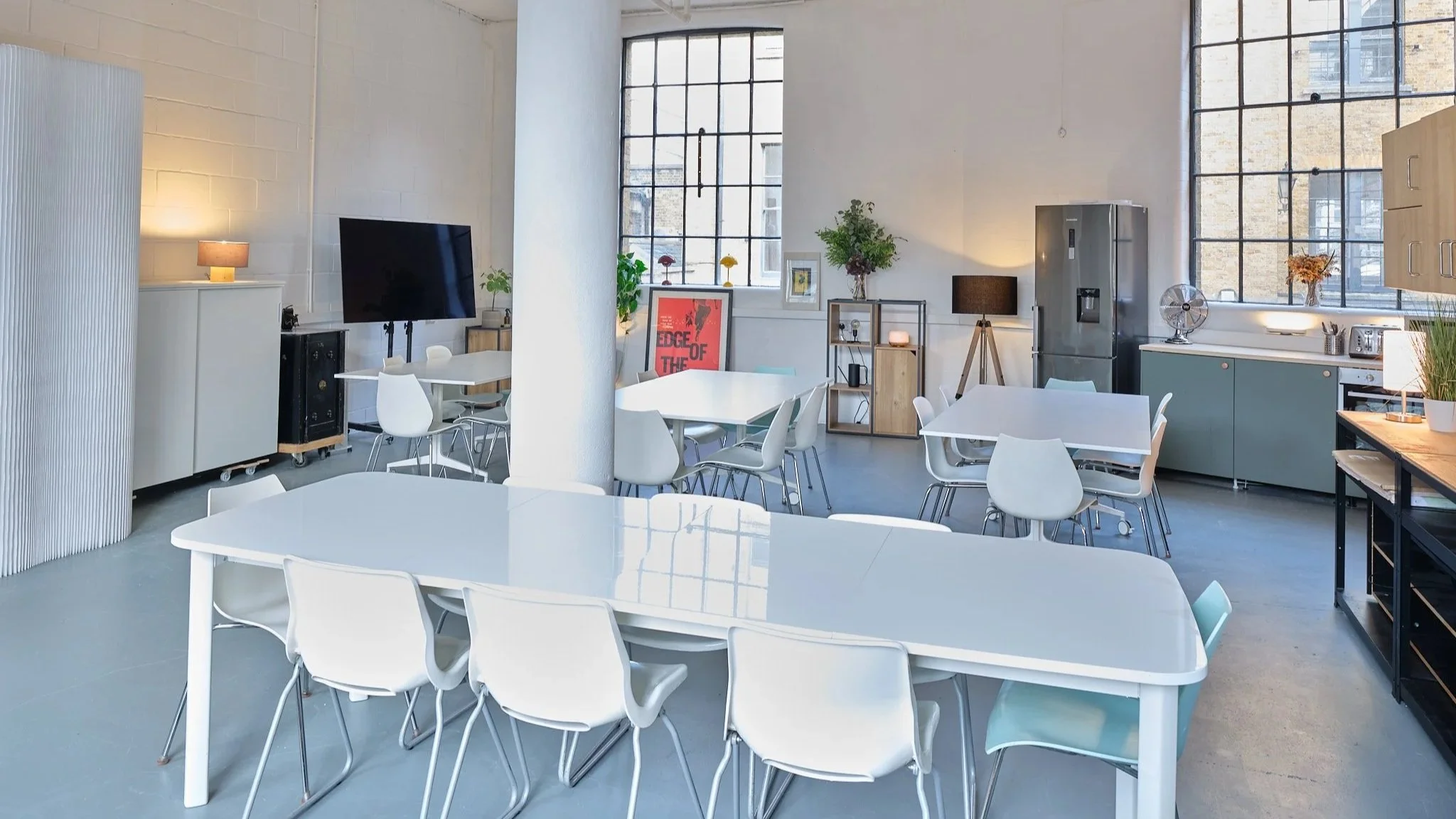 Modern kitchen and dining area with white tables, white chairs, large windows, plants, and kitchen appliances in a brightly lit loft-style space.