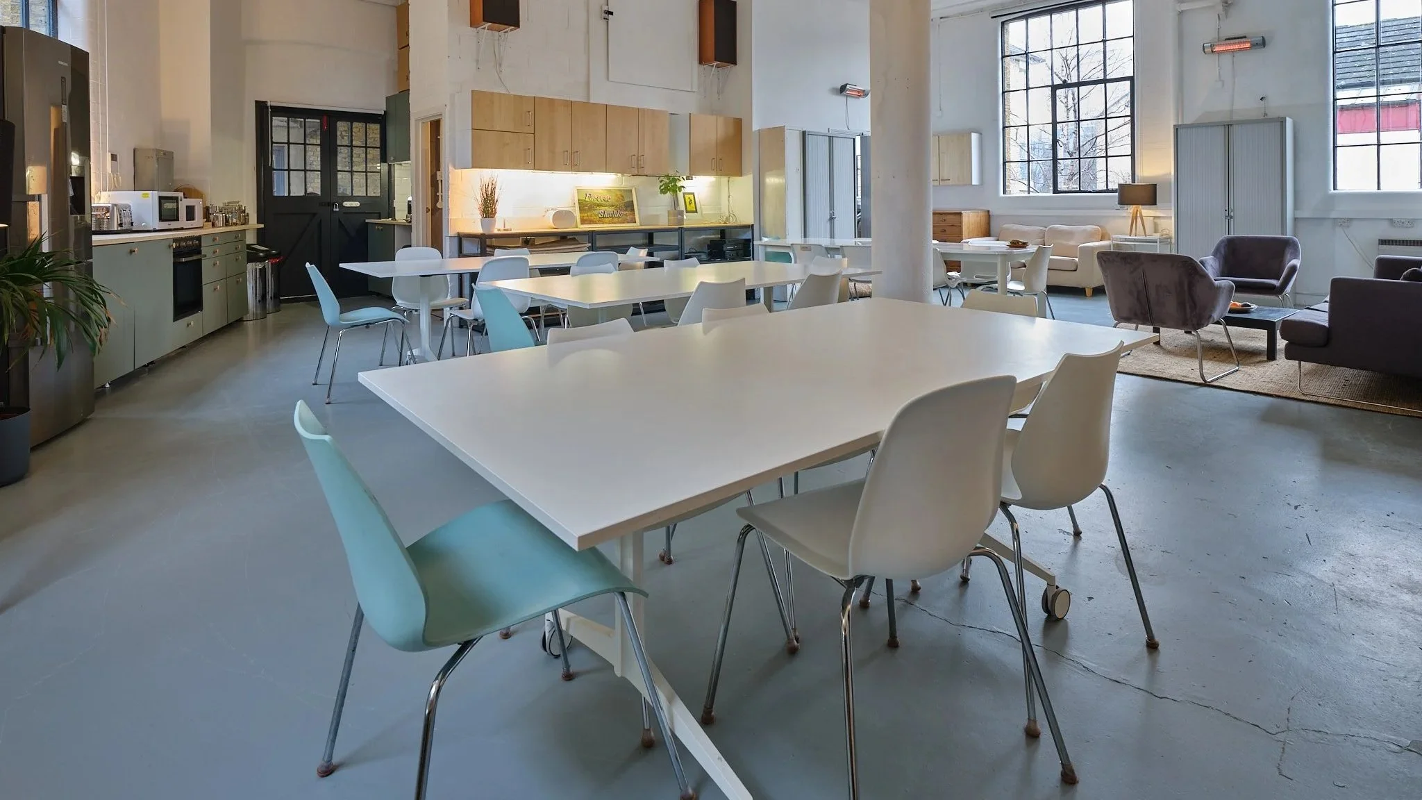 Bright, open kitchen and lounge area with white tables and assorted chairs, a kitchenette with cabinets, and a sitting area with couches and armchairs near large windows.