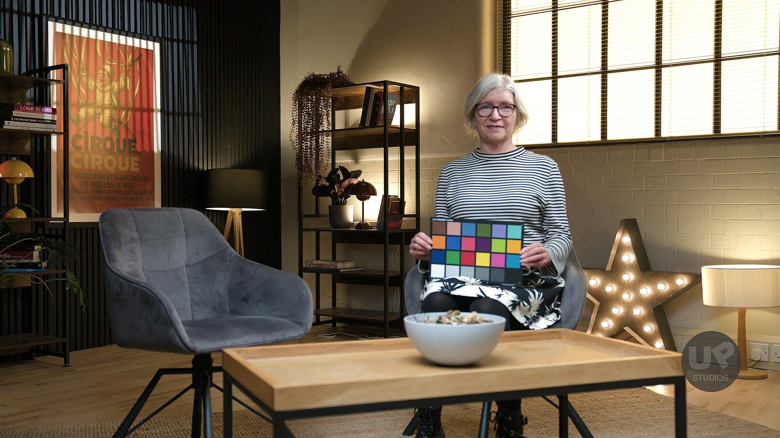 A woman with gray hair and glasses sitting in a modern living room, holding a color calibration chart. The room includes a gray chair, a wooden table with a bowl, a star-shaped light with bulbs, a bookshelf with plants and books, a lamp, and a vintag