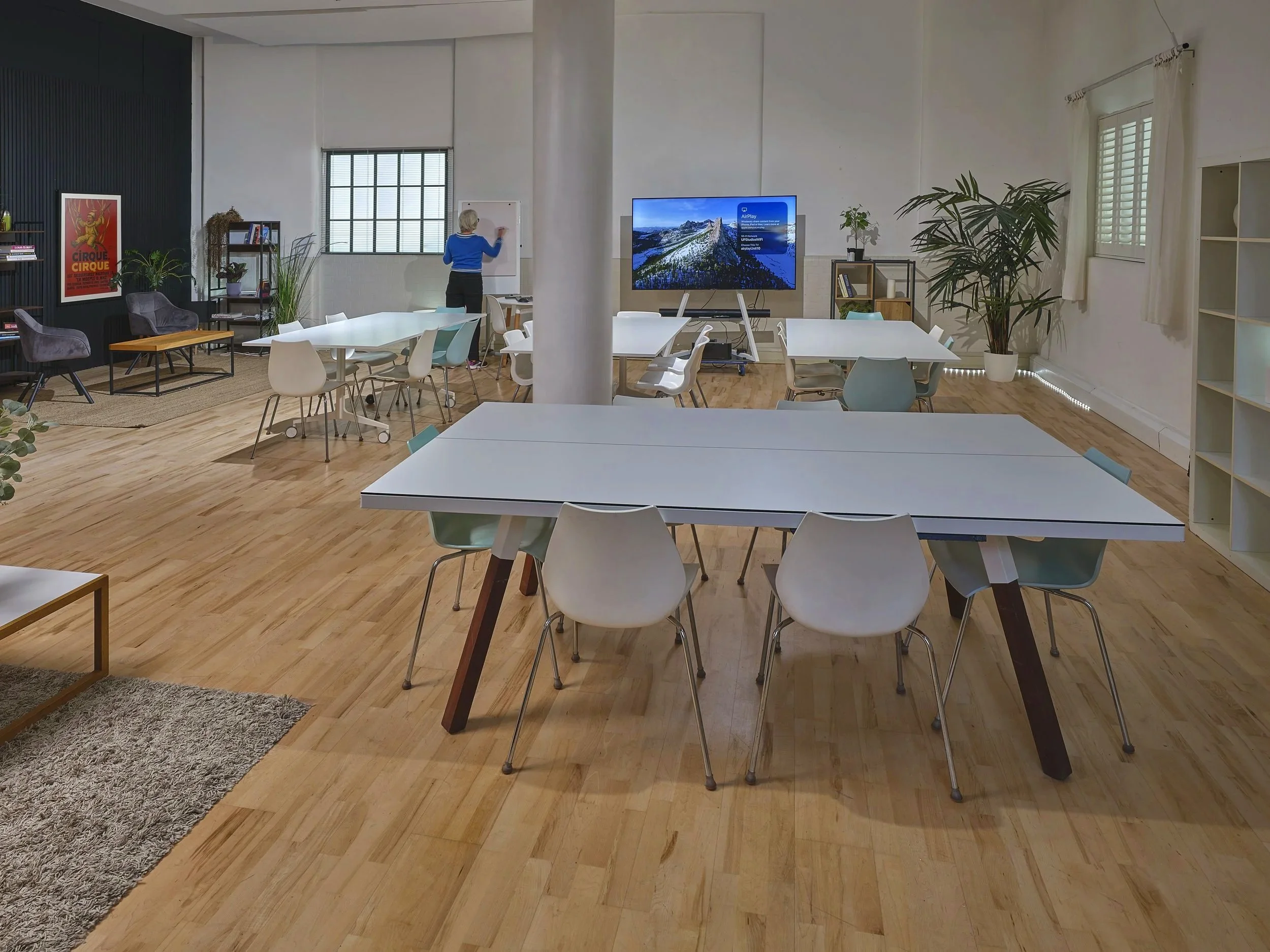 A spacious room with white tables and chairs, a person writing on a whiteboard, a large screen displaying mountain scenery, and various plants and decor.