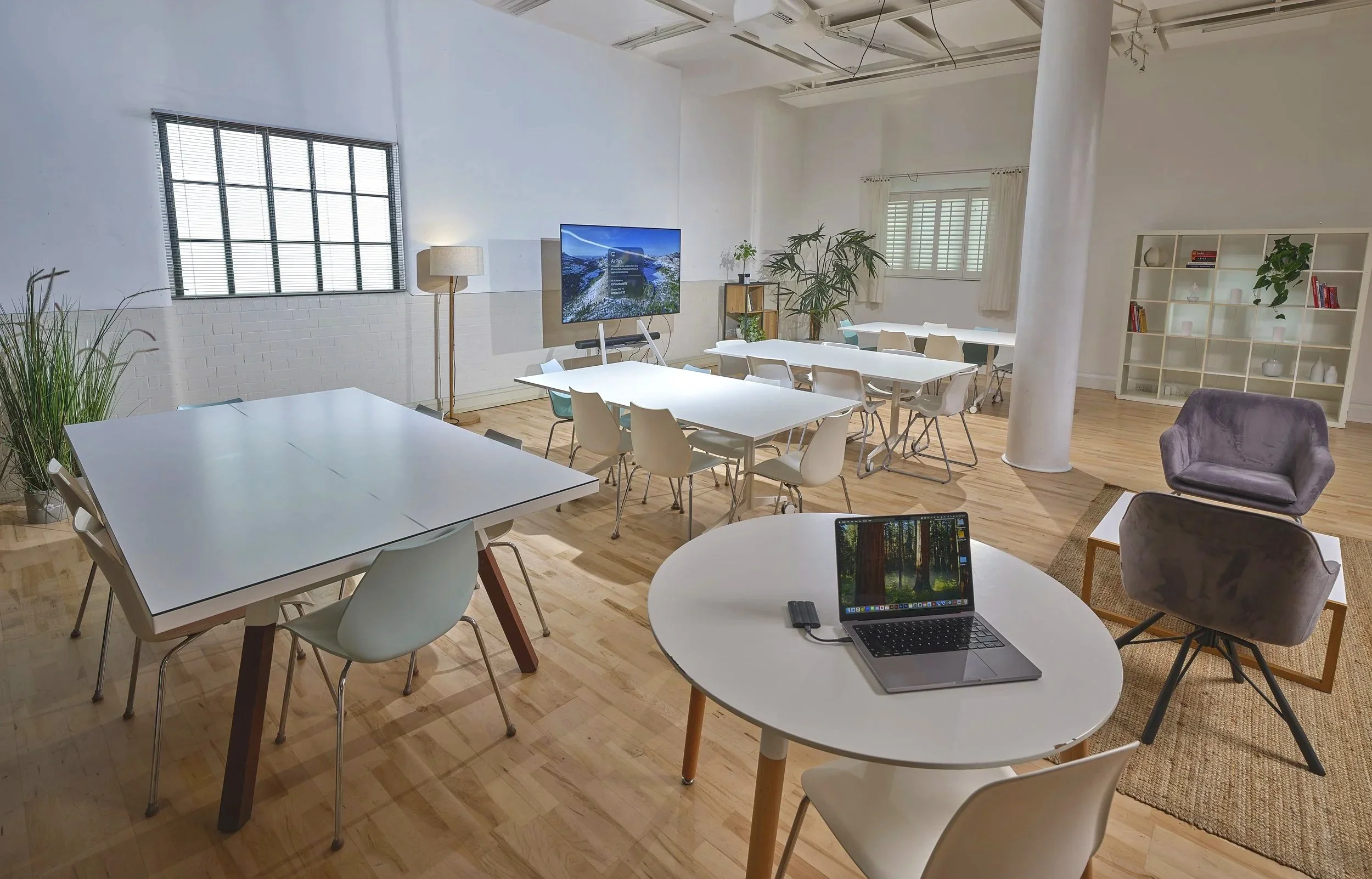 Modern conference or coworking space with white walls, light wood floors, large windows, several tables and chairs, a laptop, bookshelf, and indoor plants.