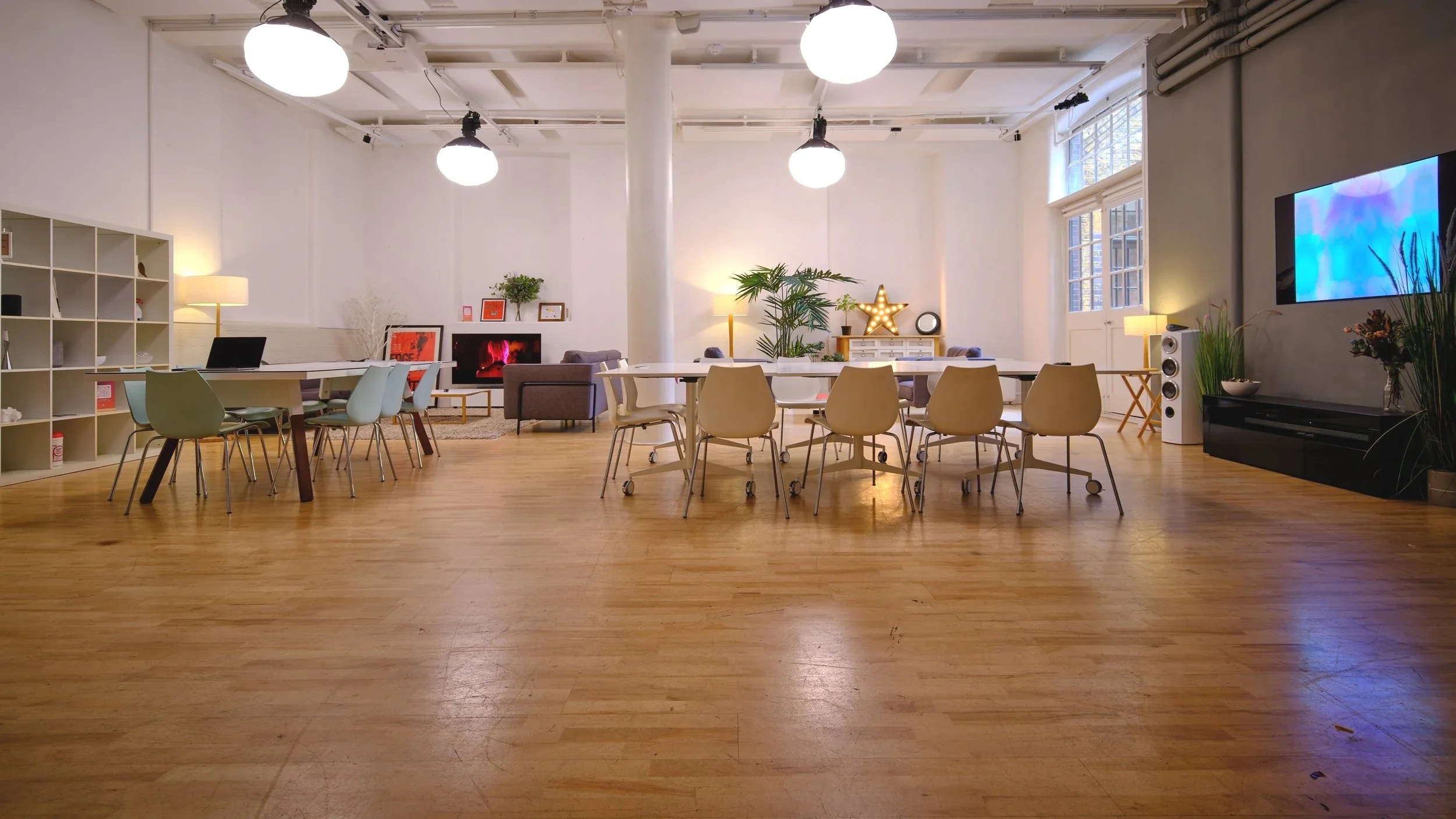 Spacious, well-lit room with a wooden floor, white walls, and large windows. It has multiple tables with chairs, a bookshelf, a wall-mounted TV, plants, and decorative items like a star-shaped light and framed artwork.