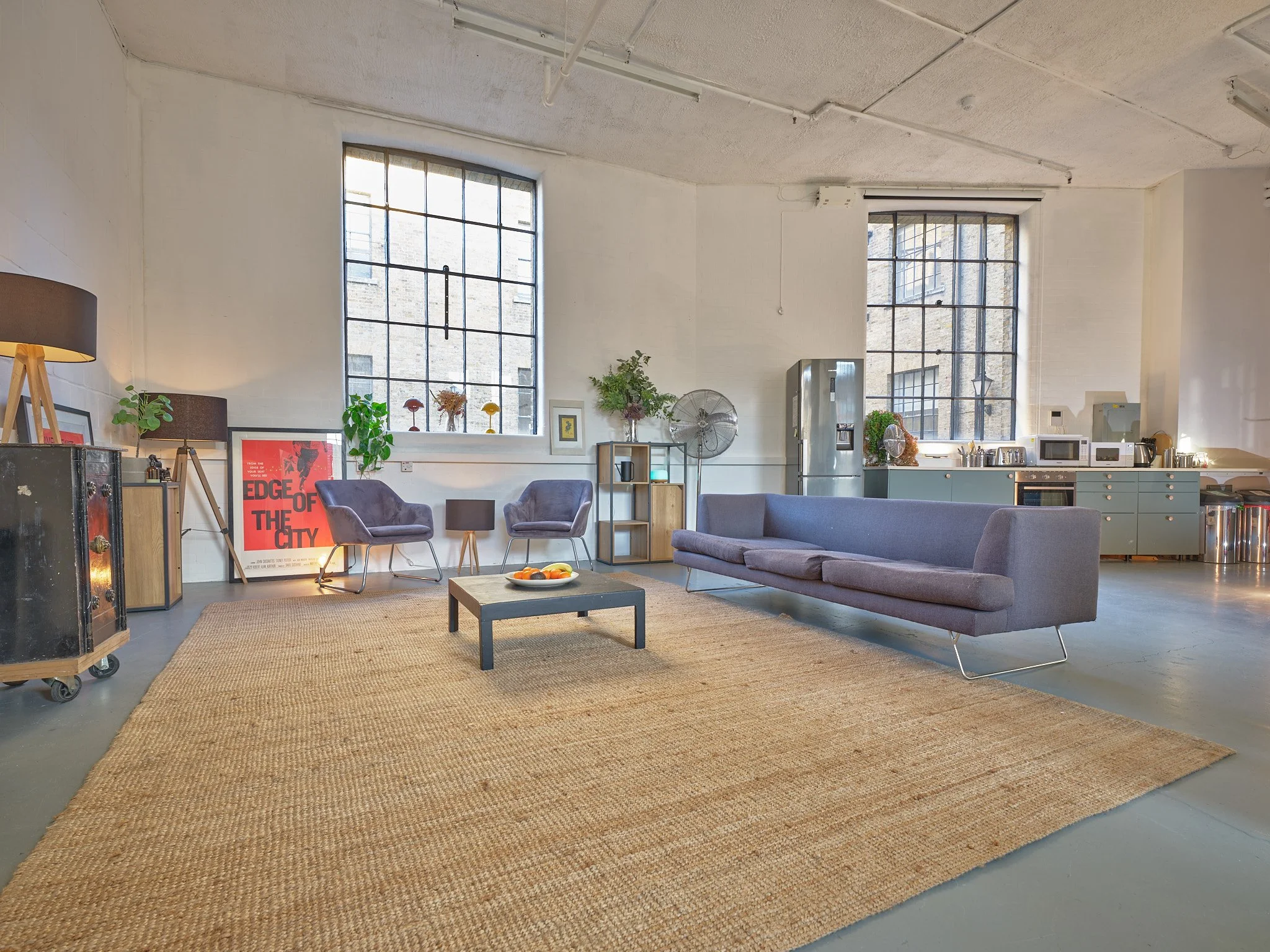 A spacious, industrial-style living room with two large windows, a grey sofa, two purple armchairs, a black coffee table with a fruit bowl, a large beige rug, and a kitchen area with pastel green cabinets and various appliances.