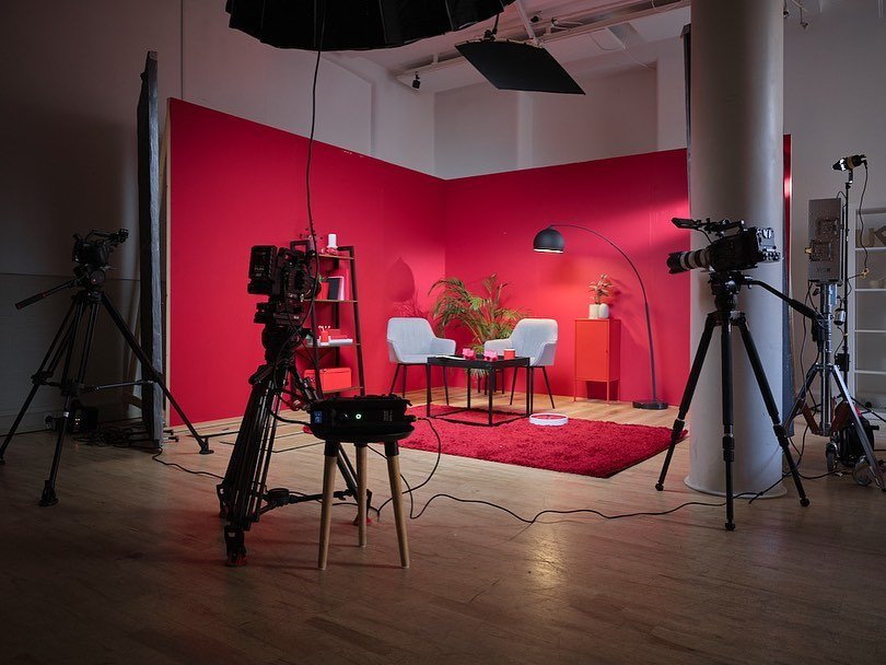 A photography studio setup with a red background, two chairs, a small table, a floor lamp, and a plant, surrounded by three professional cameras on tripods and studio lighting equipment.