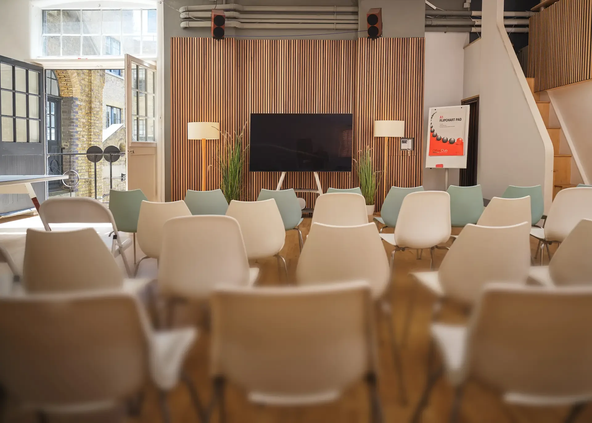 Meeting room with chairs facing a large flat-screen TV mounted on a wooden slat wall, with two tall lamps and potted plants on either side.