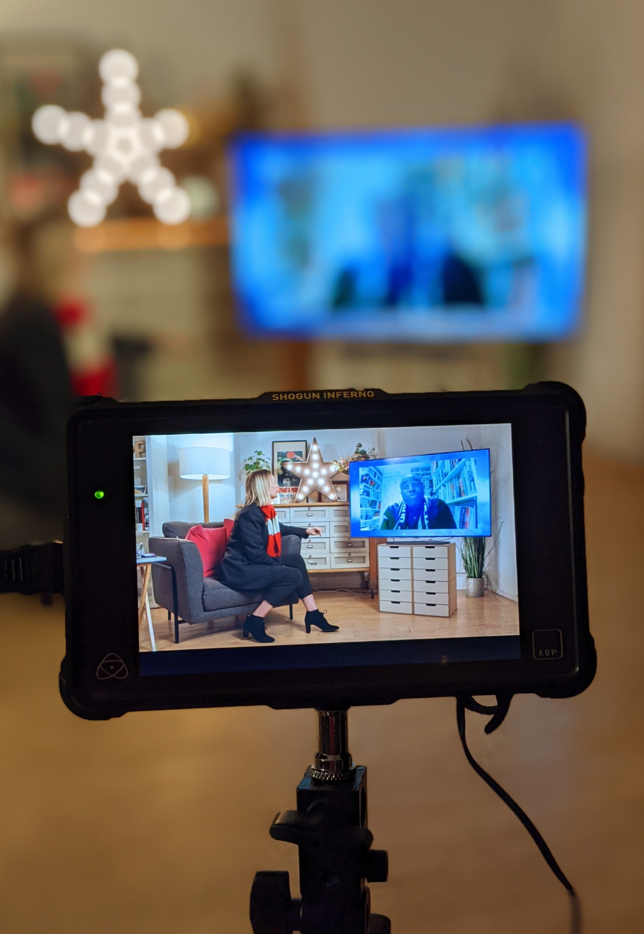 A video camera screen capturing a woman sitting on a gray couch in a decorated room, with a television screen showing the same scene in the background.