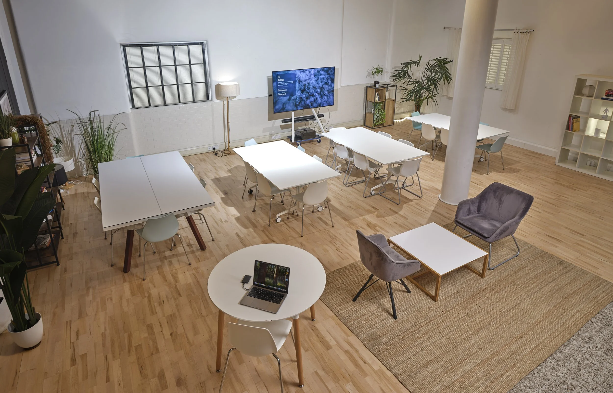 Open conference or coworking space with white tables, chairs, plants, a large screen, and a laptop on a small round table.
