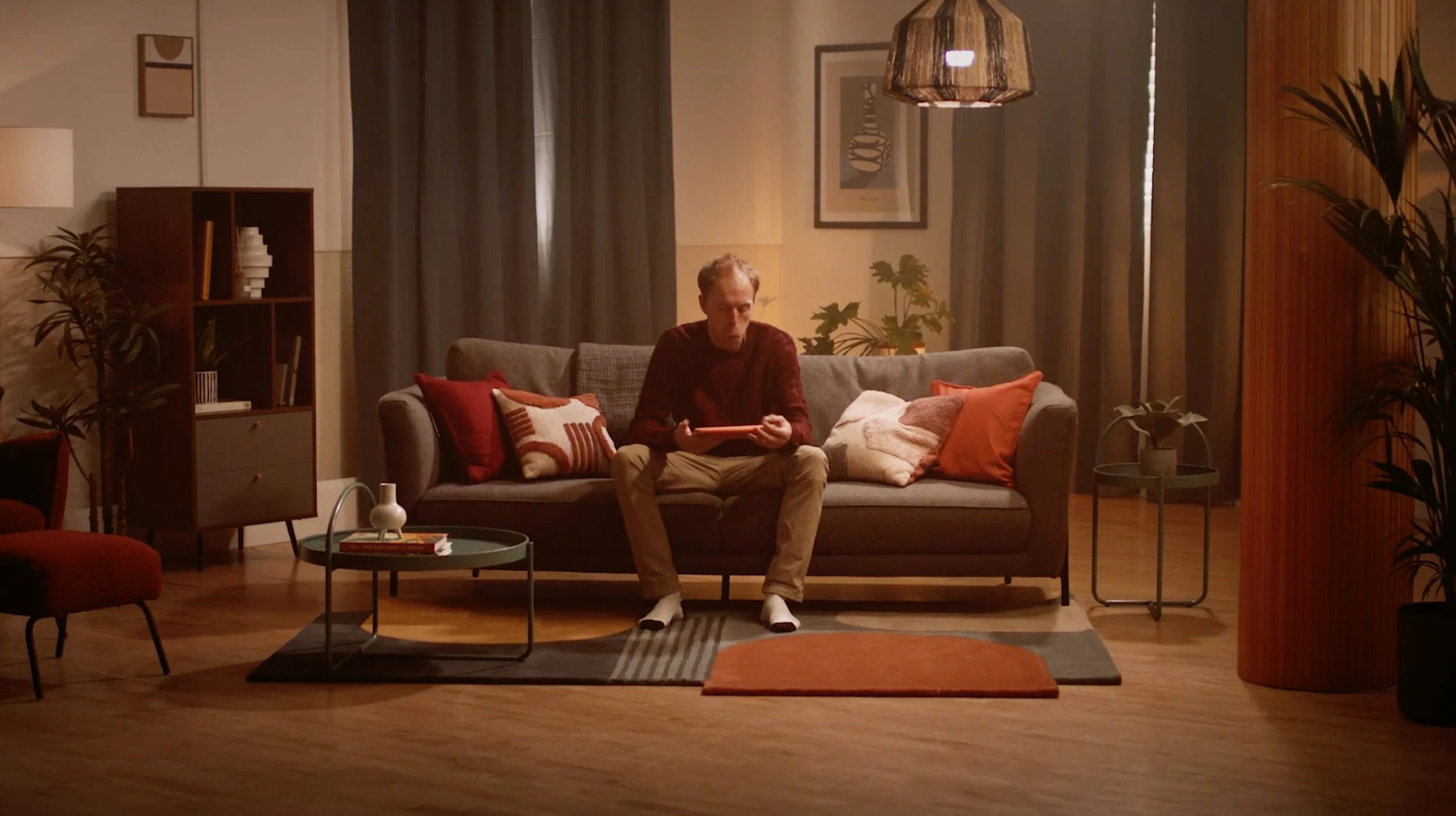 A man sitting alone on a gray sofa in a warmly lit living room, reading a tablet. The room is decorated with pillows, potted plants, a side table, a coffee table with books and a small vase, and framed art on the walls. Dark curtains cover the window