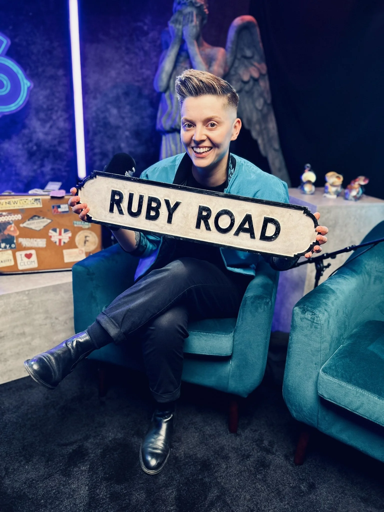 Person with short hair sitting on a teal chair, smiling, holding a 'Ruby Road' street sign, in a room with a statue of an angel and a colorful background.