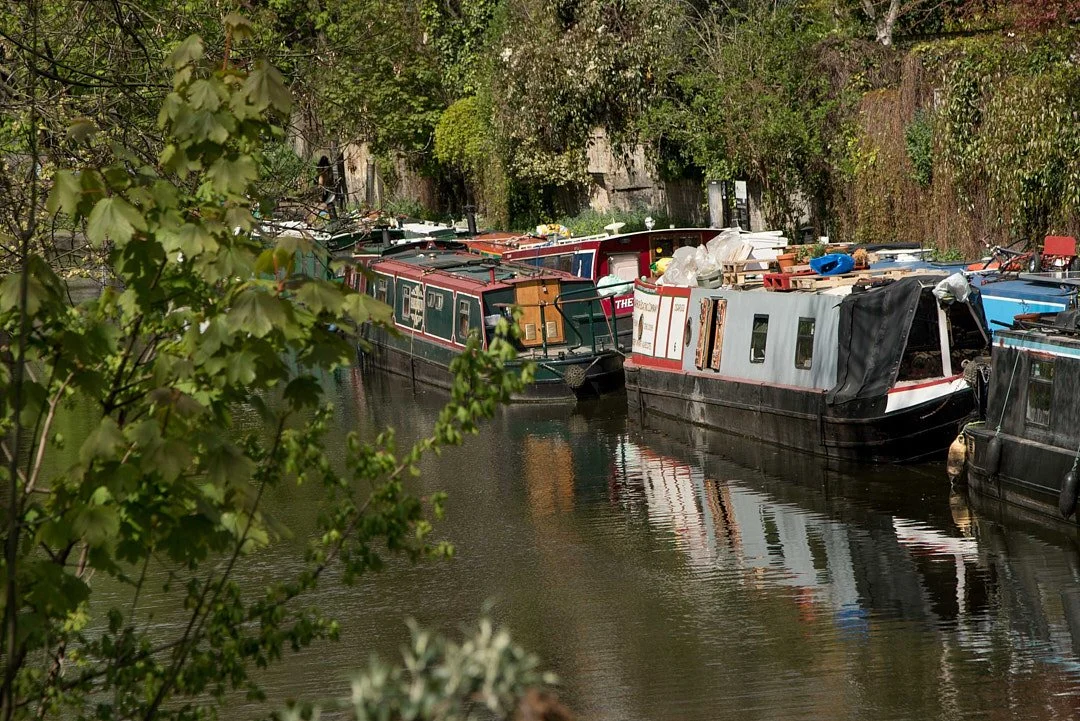 Several narrowboats docked along a narrow canal, with trees and greenery on both sides.