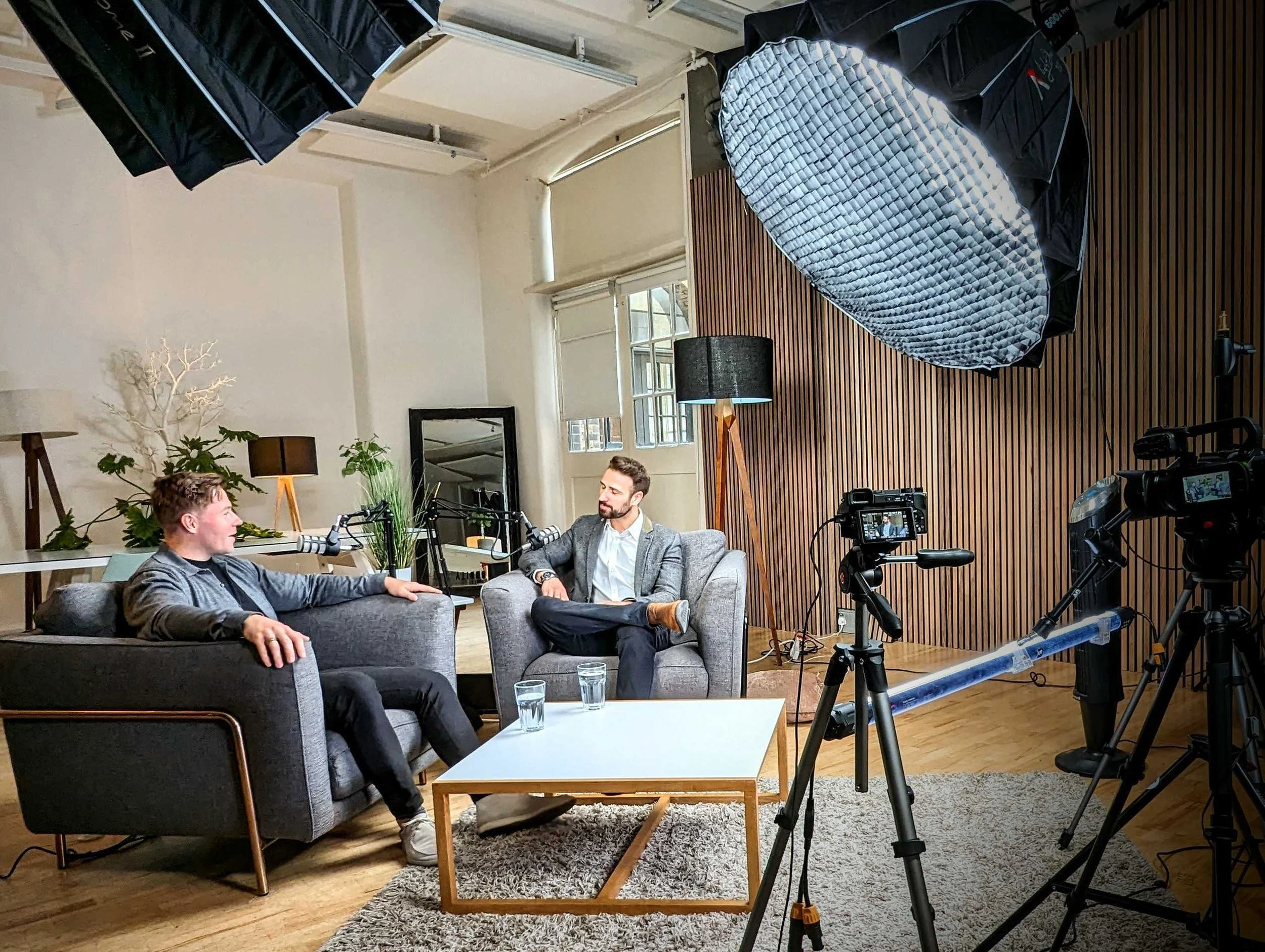 Two men having a conversation in a recording studio set up for a podcast or interview, with professional lighting and camera equipment.