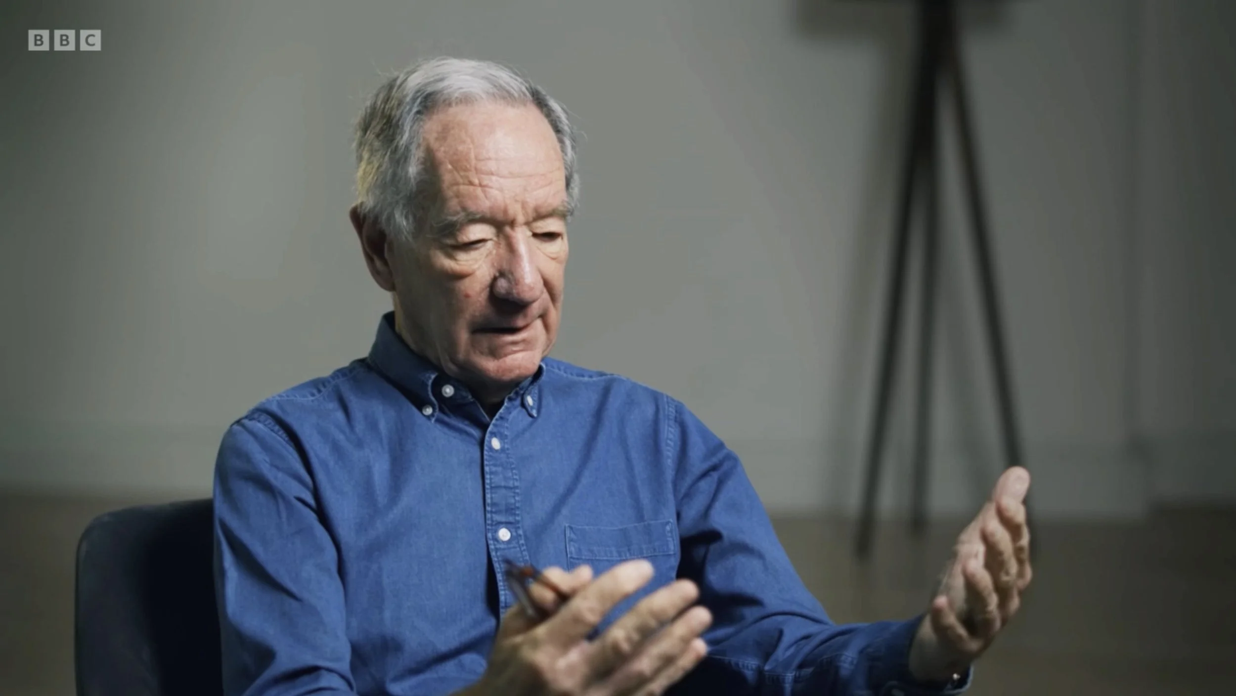 An elderly man with gray hair, wearing a blue button-up shirt, sitting in a room with a gray wall and a black tripod in the background, gesturing with his hands while speaking.