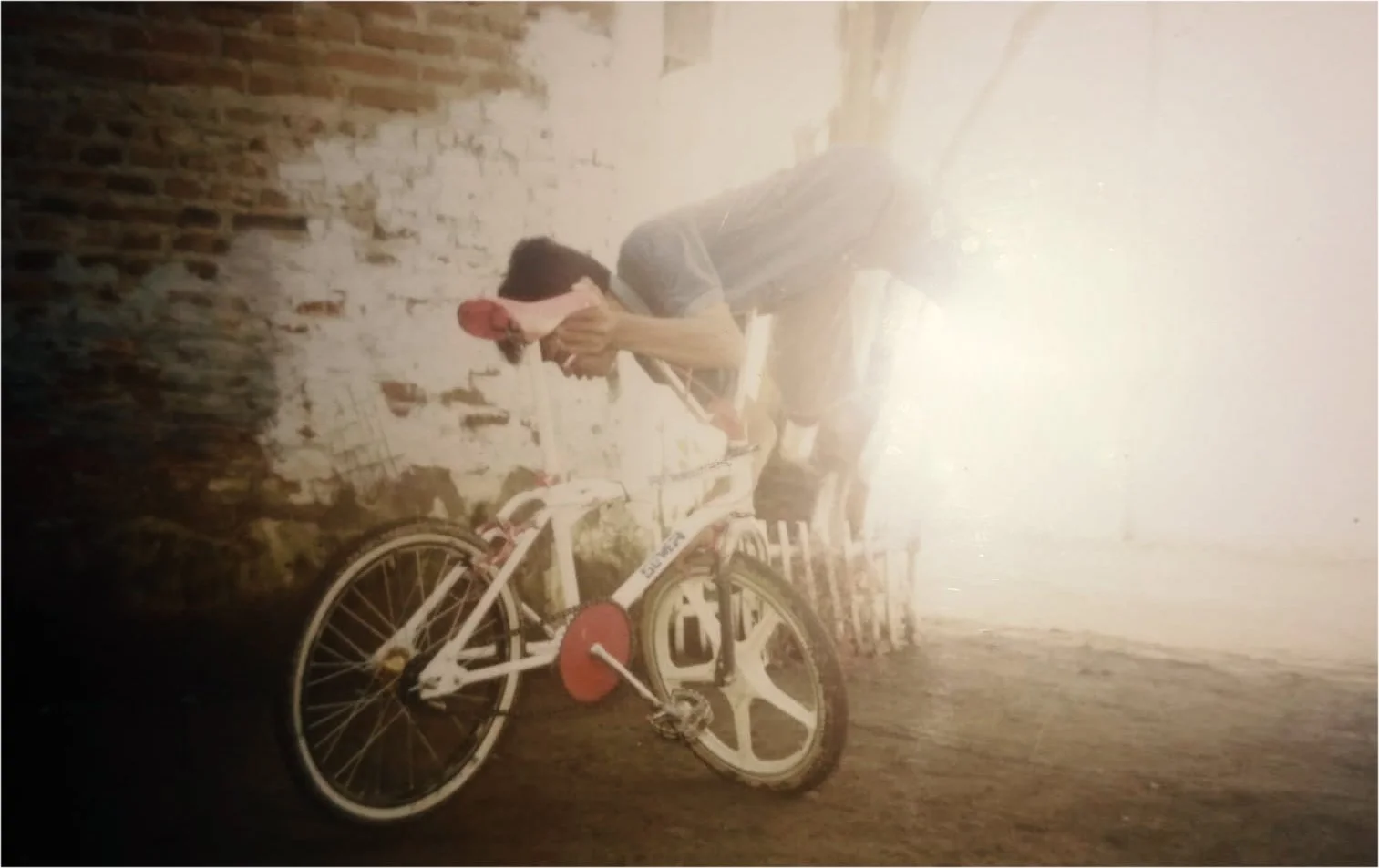 A person holding their head next to a white bicycle with red accents, standing against a rustic brick wall.