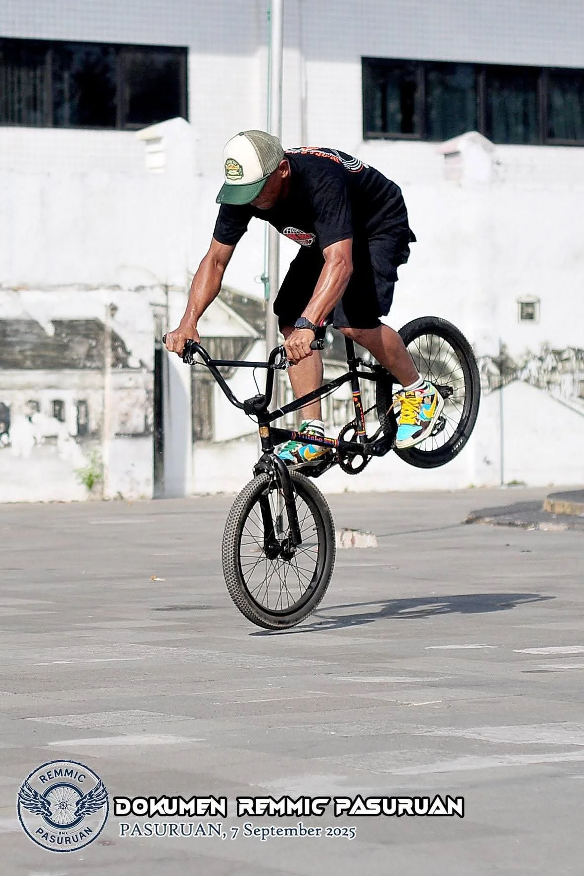 A person performing an online BMX stunt in an empty parking lot, wearing a black t-shirt, black shorts, colorful sneakers, and a baseball cap. There's a logo and text at the bottom indicating the event name, location, and date: "DOKUMEN REMMIC PASURUAN, PASURUAN, 7 September 2025."