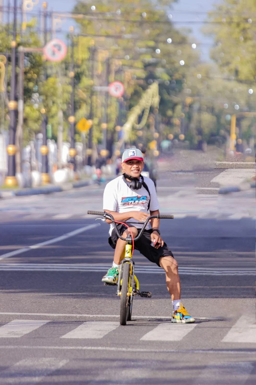 A man is riding a yellow bicycle in the middle of a street, smiling, with one foot on the pedal and the other on the ground. He is wearing a white T-shirt, black shorts, a baseball cap, and colorful sneakers, with headphones around his neck.