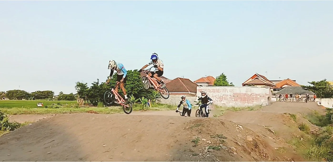 Children riding BMX bikes and playing outdoors on a dirt track with houses and trees in the background.