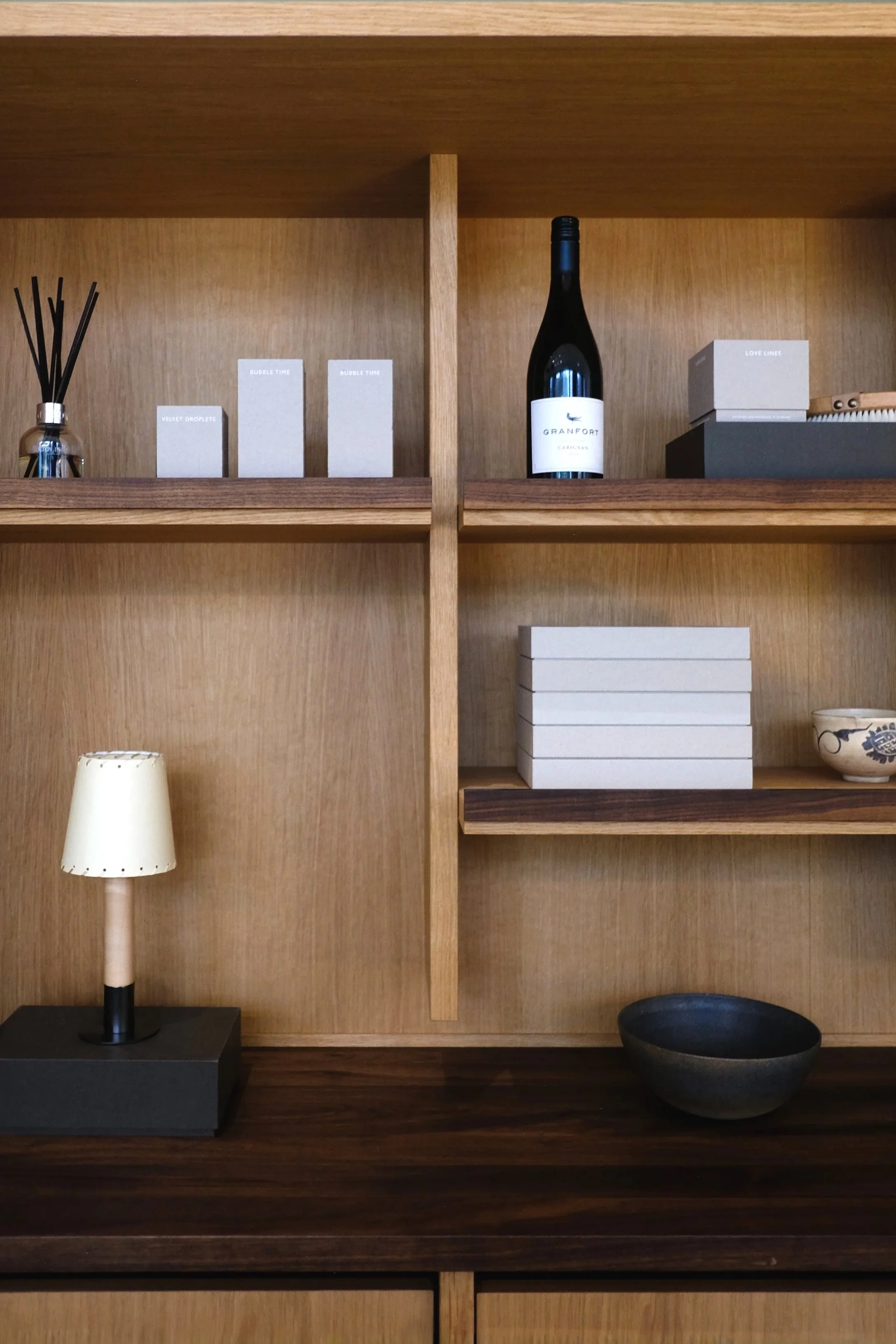 Wooden bookshelf with decorative items including a small lamp, stack of white boxes, scented diffuser, and a bottle of wine.