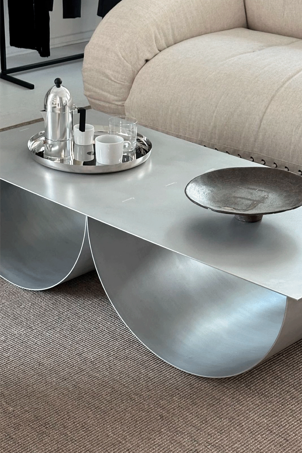 Modern silver coffee table with a curved base, holding a silver tray with a coffee press, cups, and glasses, placed near a beige sofa.