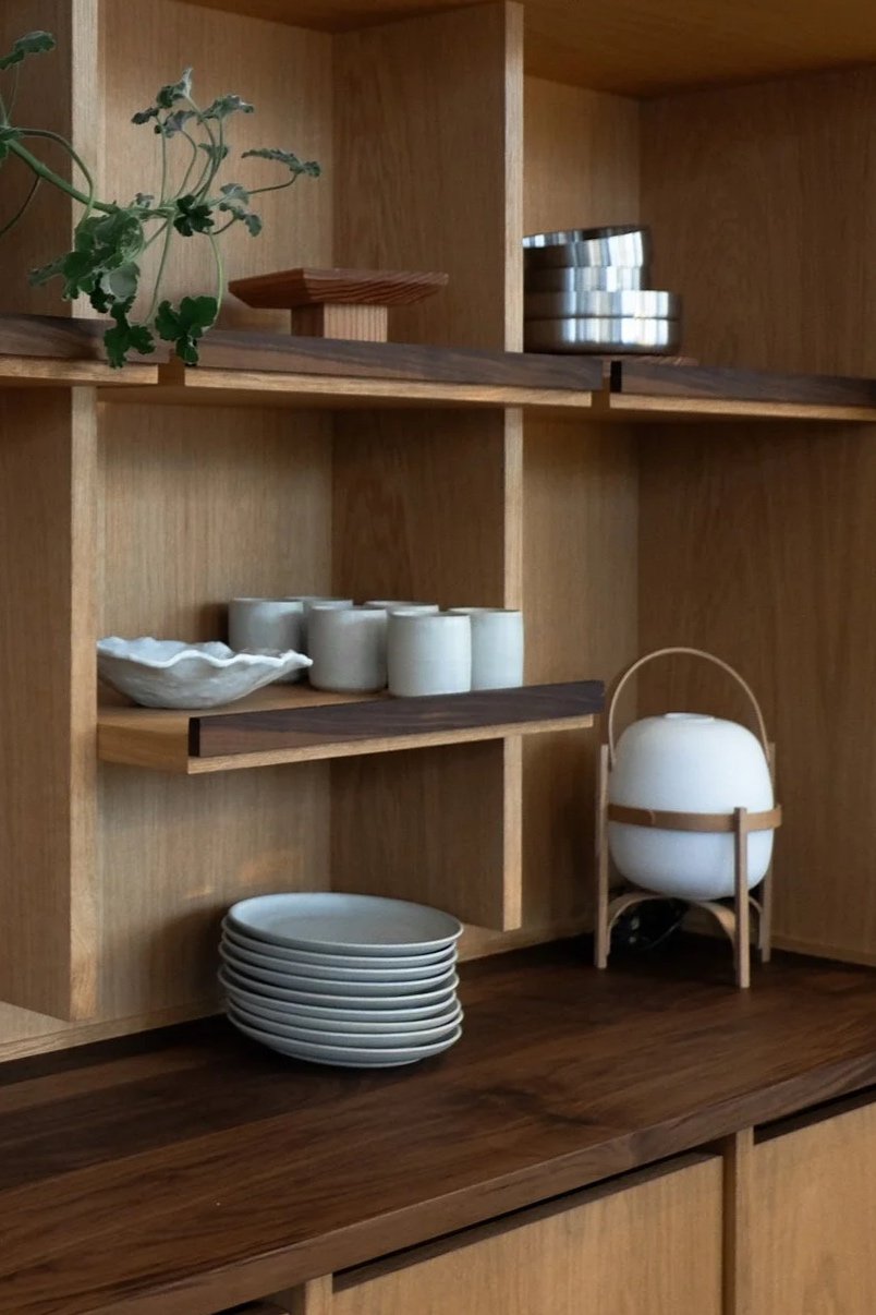 Wooden shelves holding white plates, cups, small bowls, a decorative white vessel with a wooden handle, and a small plant in a white pot.