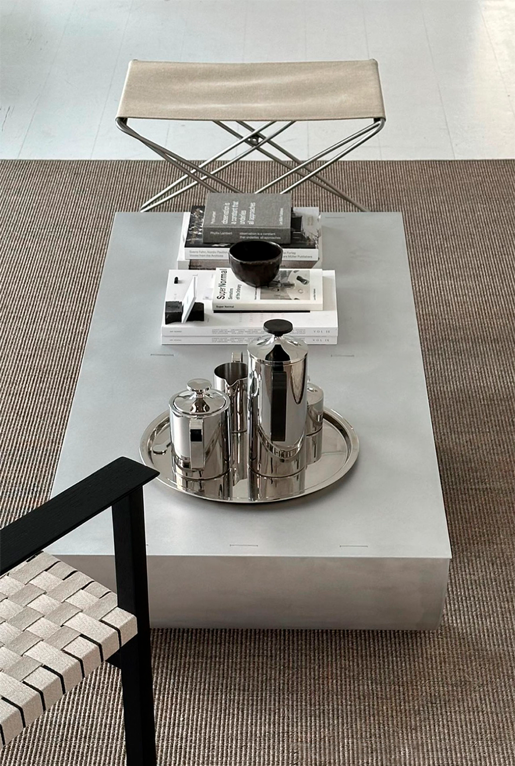 Top view of a modern coffee table with books, a black bowl, and a silver tea set on a tray, with a beige bench behind it and a black and white chair to the side.
