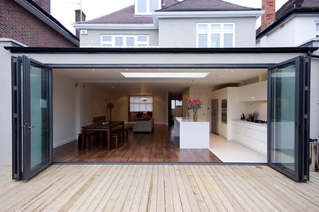 A modern house with large open folding glass doors leading to a wooden deck, revealing an indoor living space with a white kitchen island with a vase of pink flowers, a dining area, and a sitting area in the background.