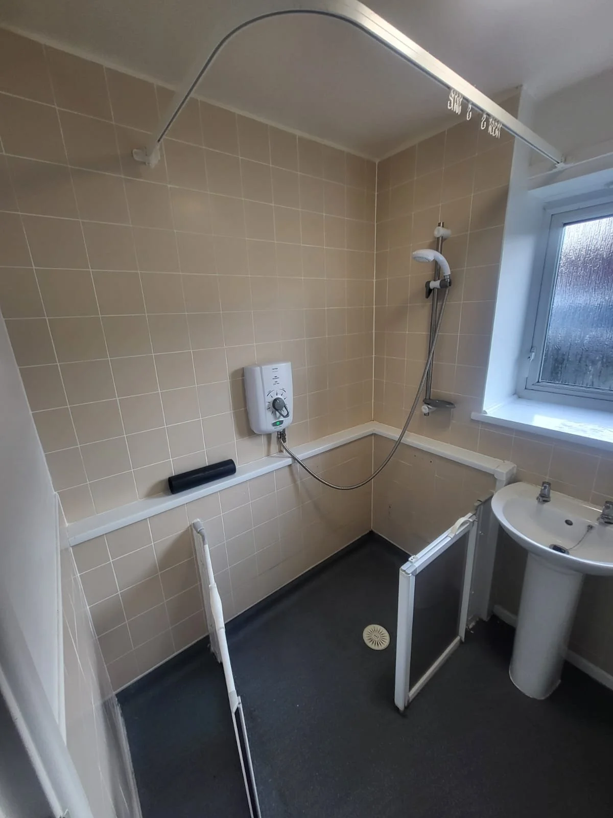 A shower room with beige tiled walls, a showerhead connected to an electric water heater, a small sink with two faucets, a window, and a folding shower screen.