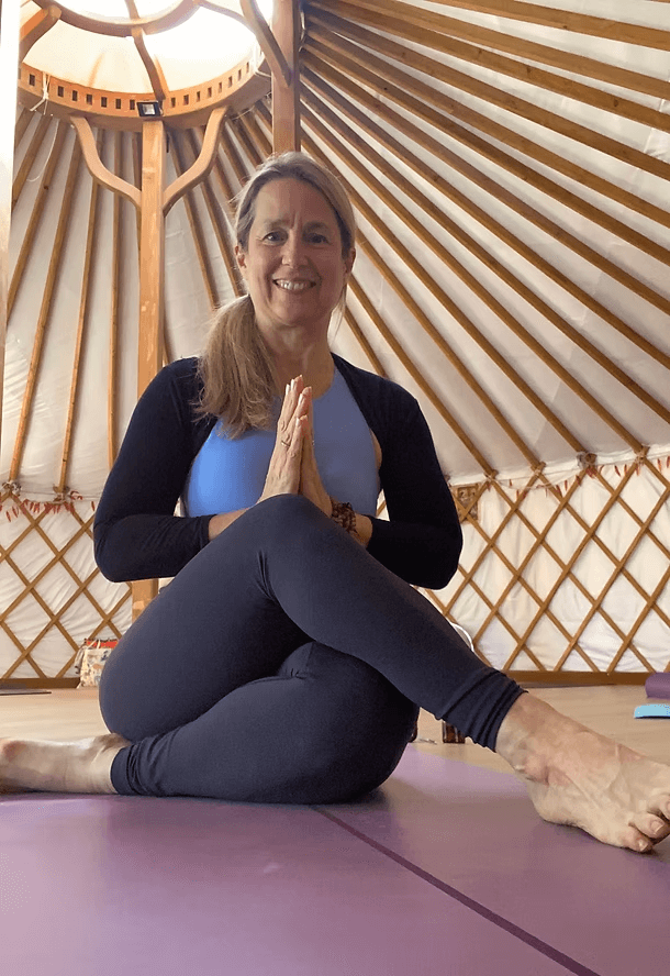 A woman practicing yoga inside a yurt, sitting cross-legged with palms pressed together in a prayer position, smiling at the camera.