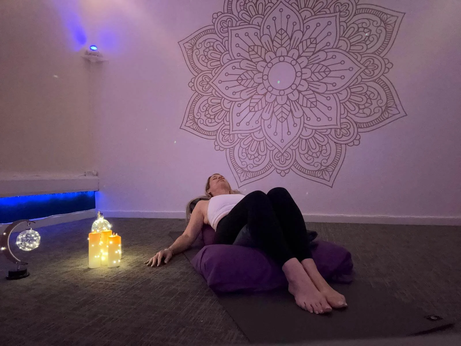 Woman lying on her back on a yoga mat with a purple pillow under her head, inside a dimly lit room with a large decorative mandala pattern on the wall, candles, and decorative lights.