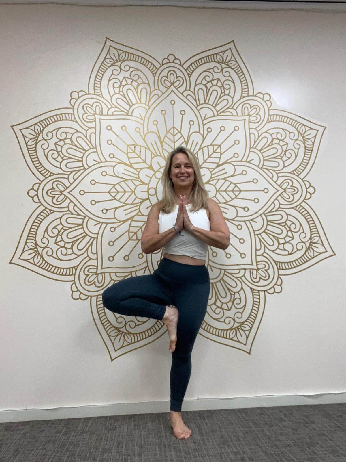 A woman practicing yoga indoors, standing on one leg in the tree pose, with hands in a prayer position, in front of a large, decorative mandala wall art.